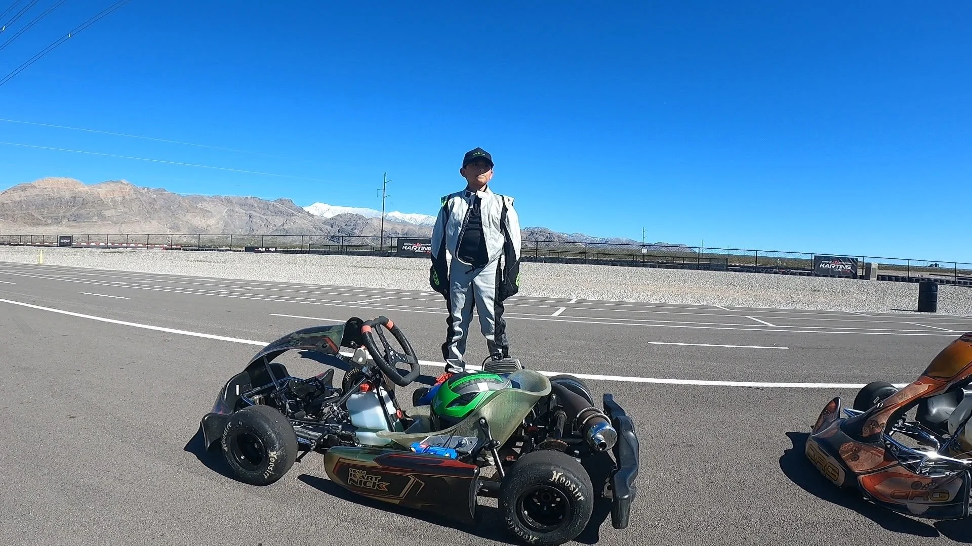 A young boy in racing gear stands on a race track with remote control go-karts in front of him, with mountains in the background under a clear blue sky.
