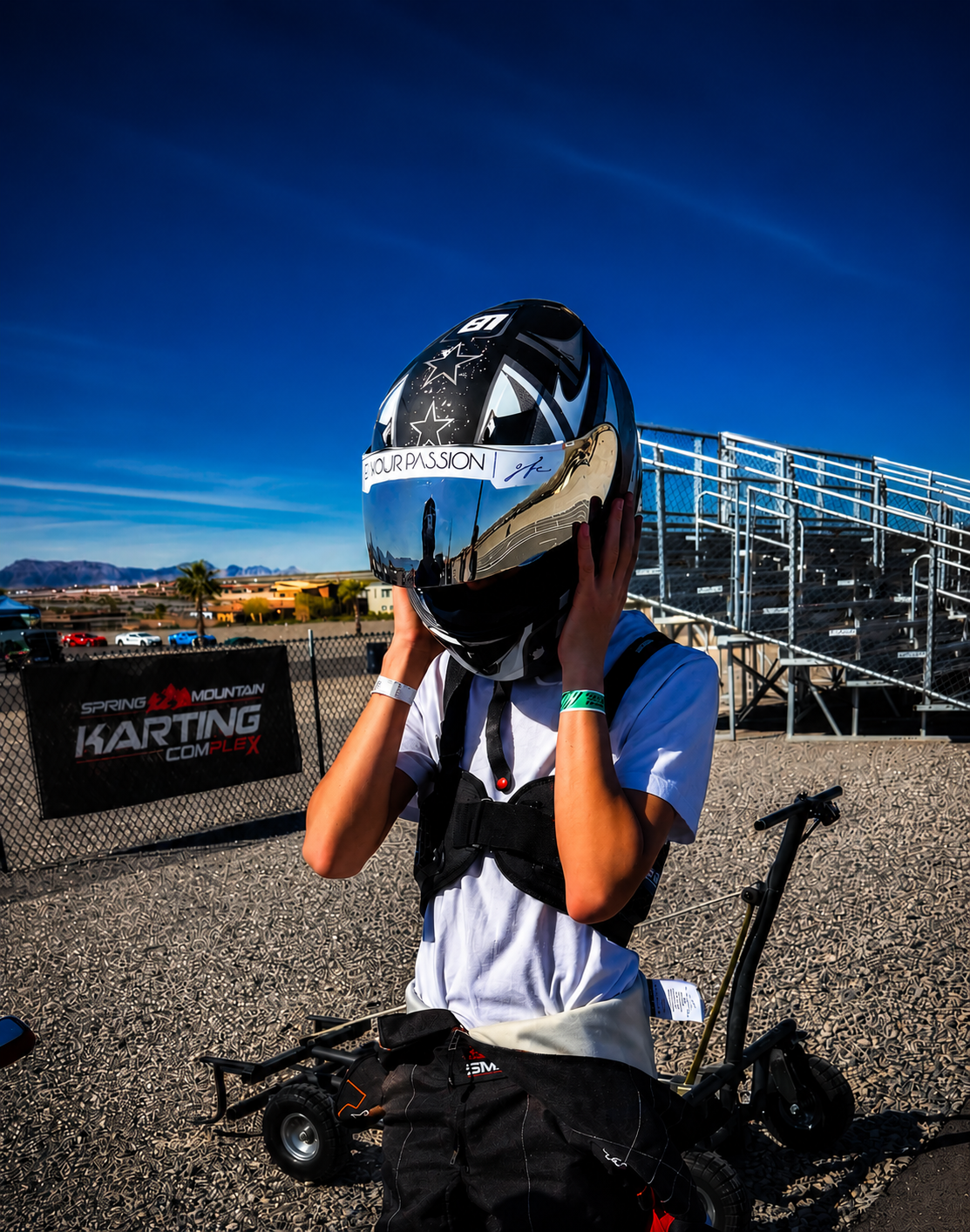 Person in a white race suit adjusting a racing helmet at a go-kart track, with a sign reading 'Spring Mountain Karting Complex' and bleachers in the background under a clear blue sky.