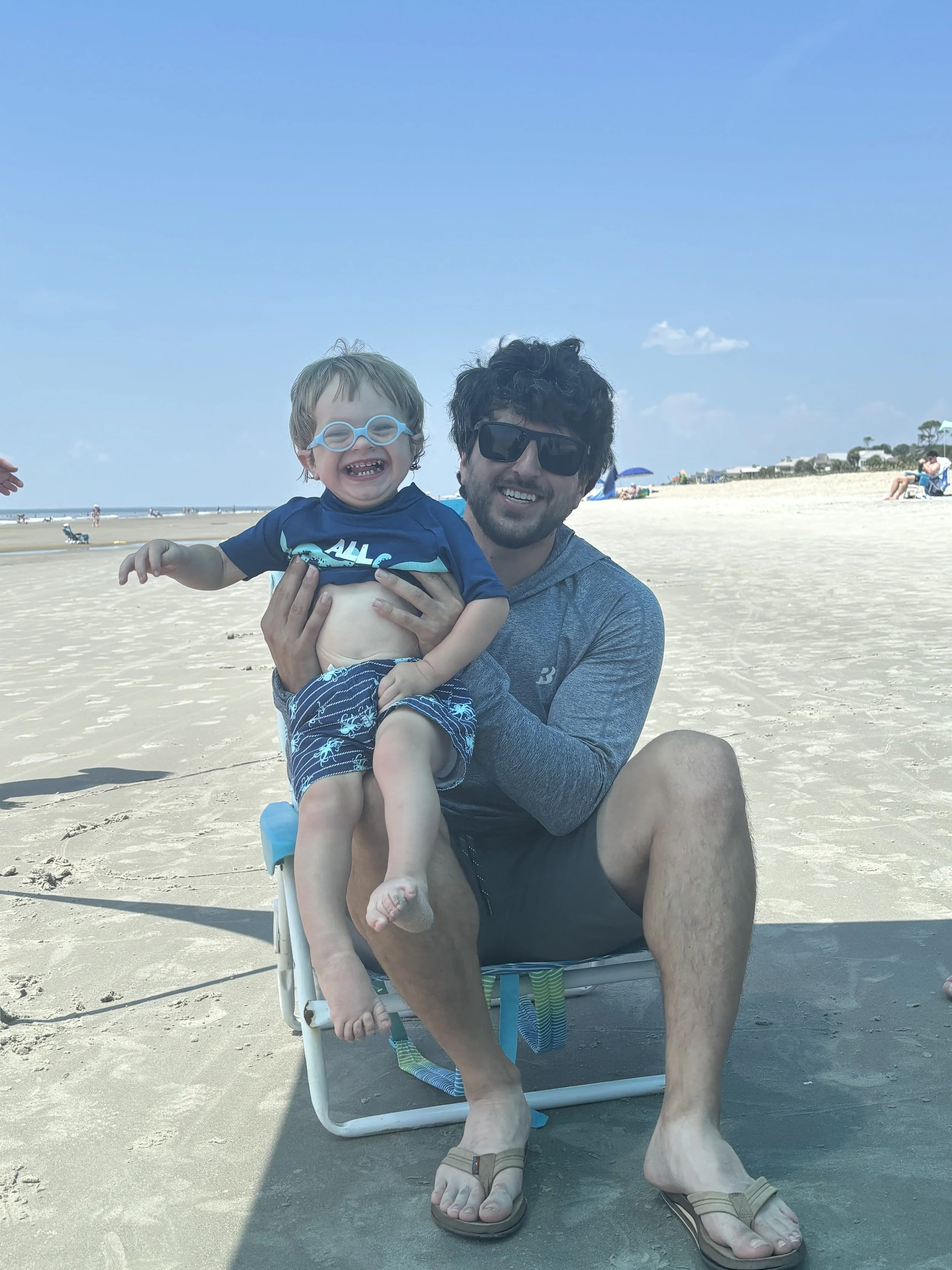 A man and a young boy at the beach, smiling and enjoying their time. The boy is wearing swimming goggles, a blue shirt, and patterned shorts. The man is wearing sunglasses, a gray hoodie, and black shorts. They are sitting on a beach chair on the sand, with a clear blue sky and some people in the background.