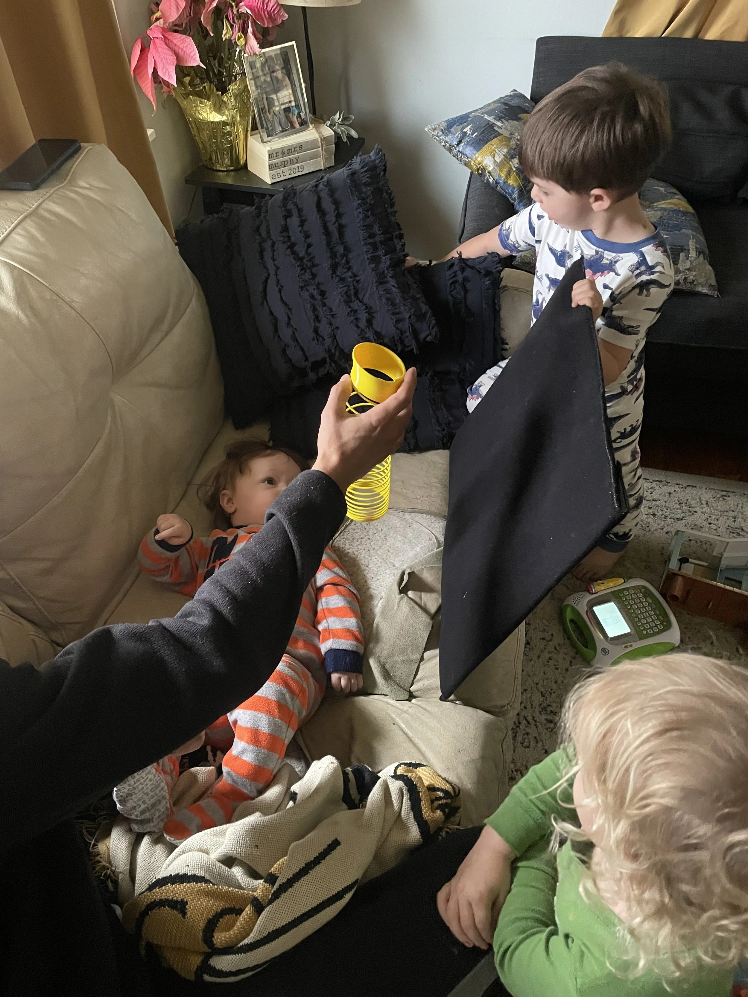 Baby in orange striped footie pajamas lays on couch and looks at black board. Long arm holds yellow slinky in field of vision, backdropped by the black board