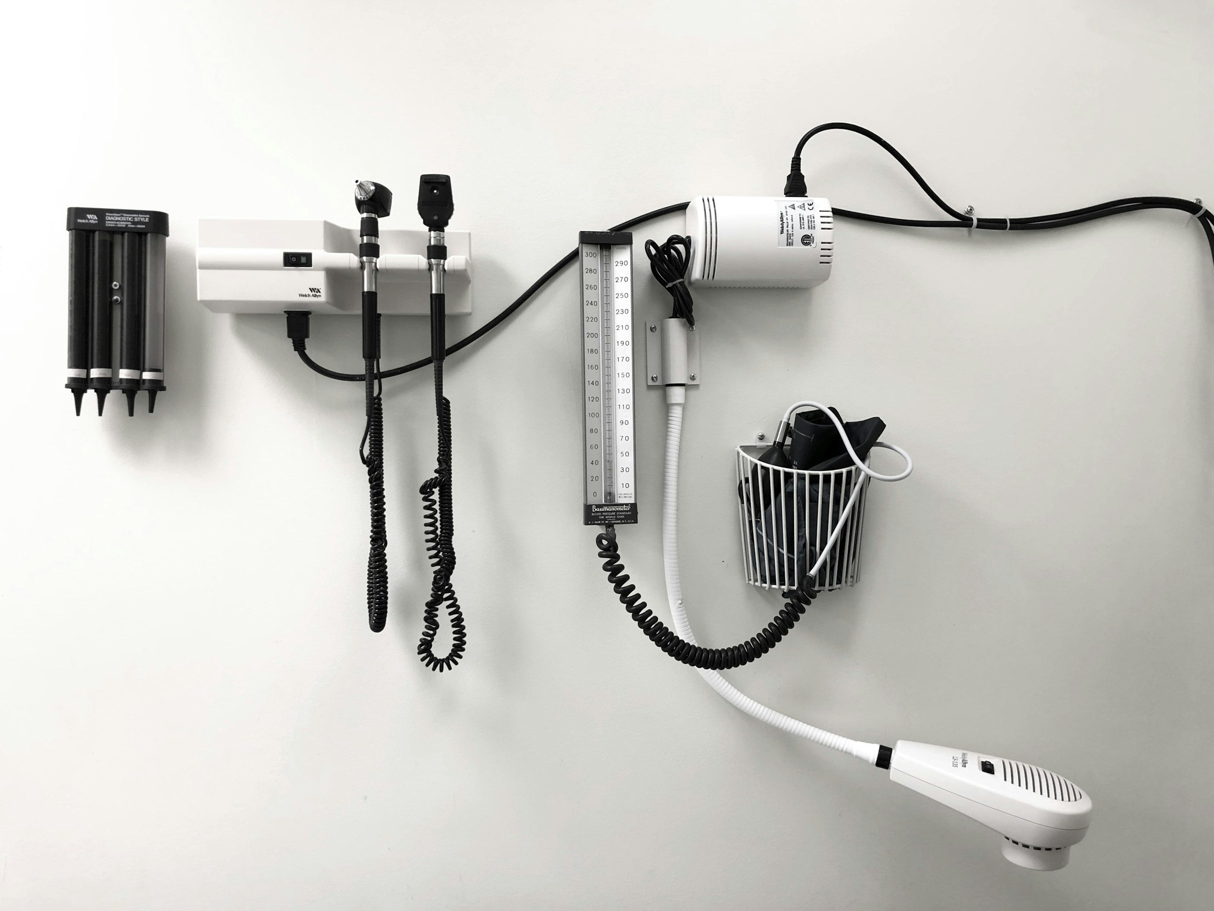 Medical equipment on a white wall, including a black diagnostic device, two probe attachments, a white monitor, a pressure gauge, and various cords and accessories in a wire basket.