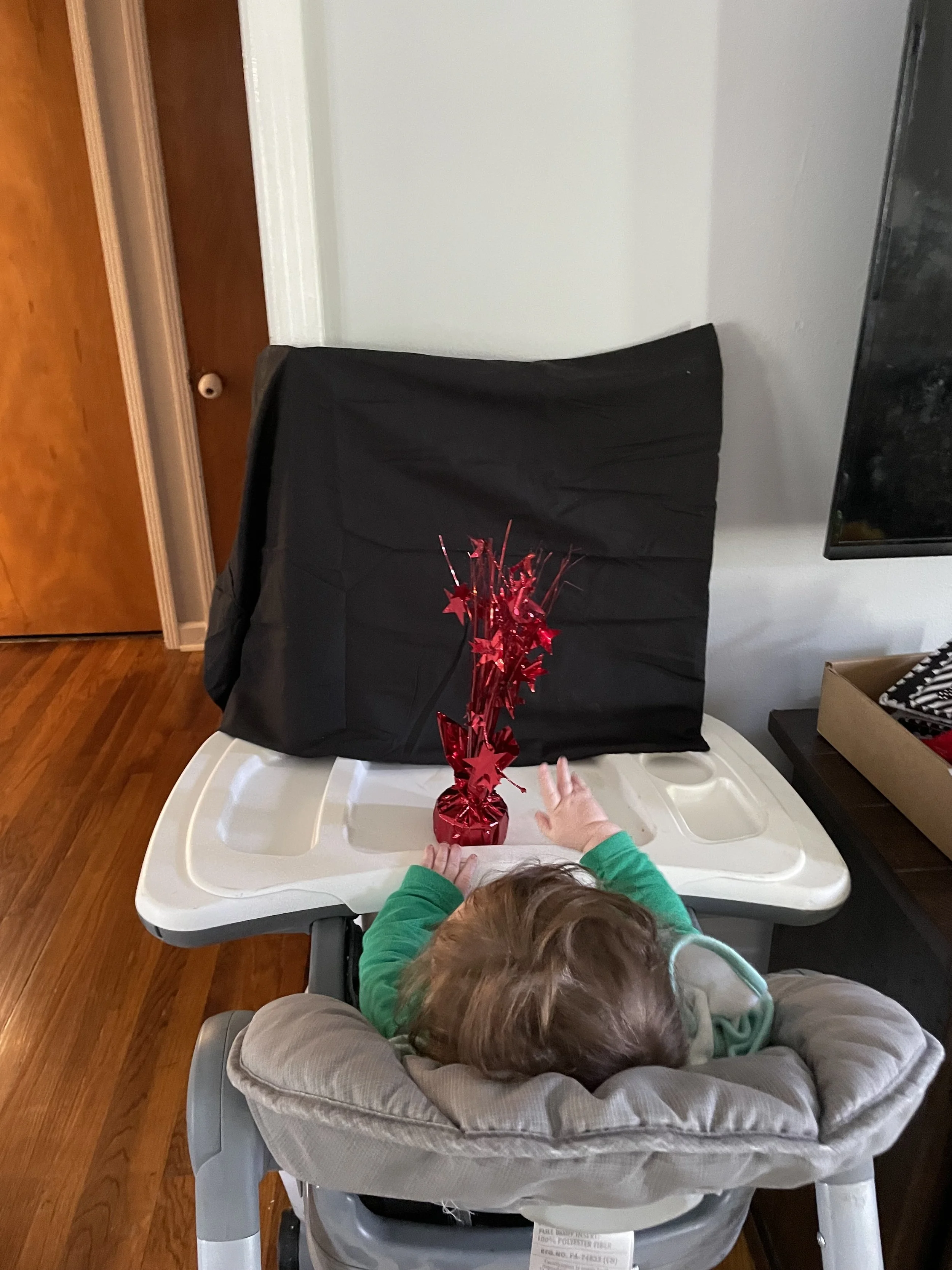 Baby sits in high chair facing a wall. There is a black board in front of him with a shiny red table decoration. 