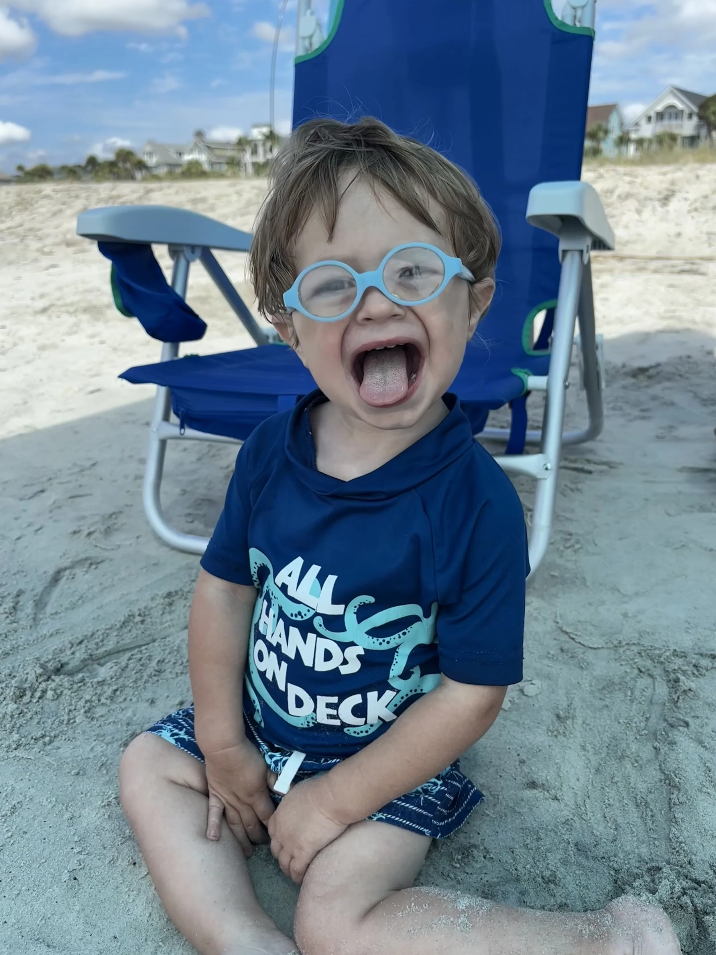 A young boy is sitting on the sandy beach, wearing blue glasses and a dark blue t-shirt with white text, sticking out his tongue. A blue beach chair is behind him, and there are houses and a blue sky in the background.