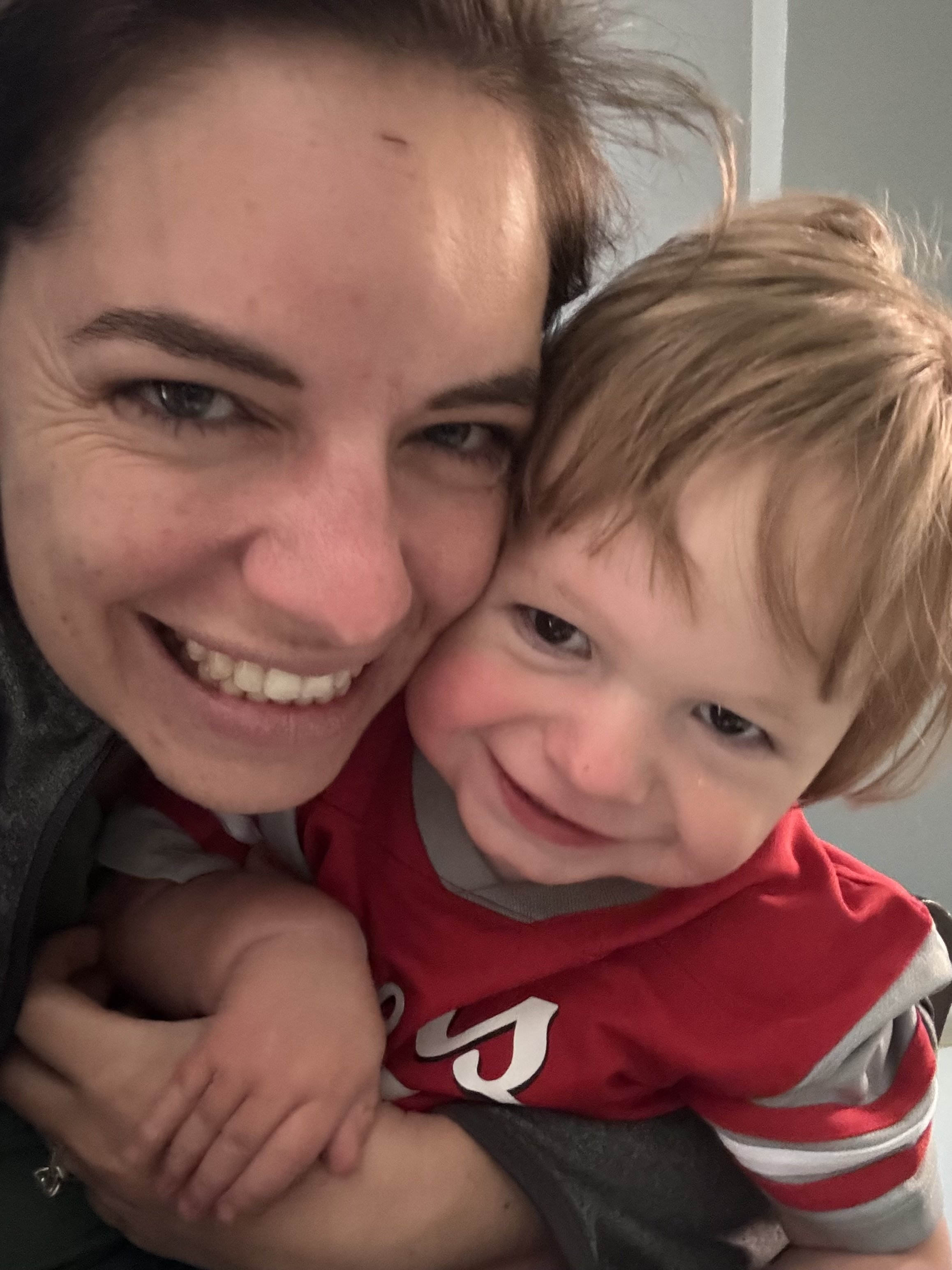 A woman and a young boy are close together, smiling and looking at the camera. The woman has dark hair and is wearing a dark top, while the boy has light brown hair and is wearing a red sports jersey. They appear happy and affectionate.