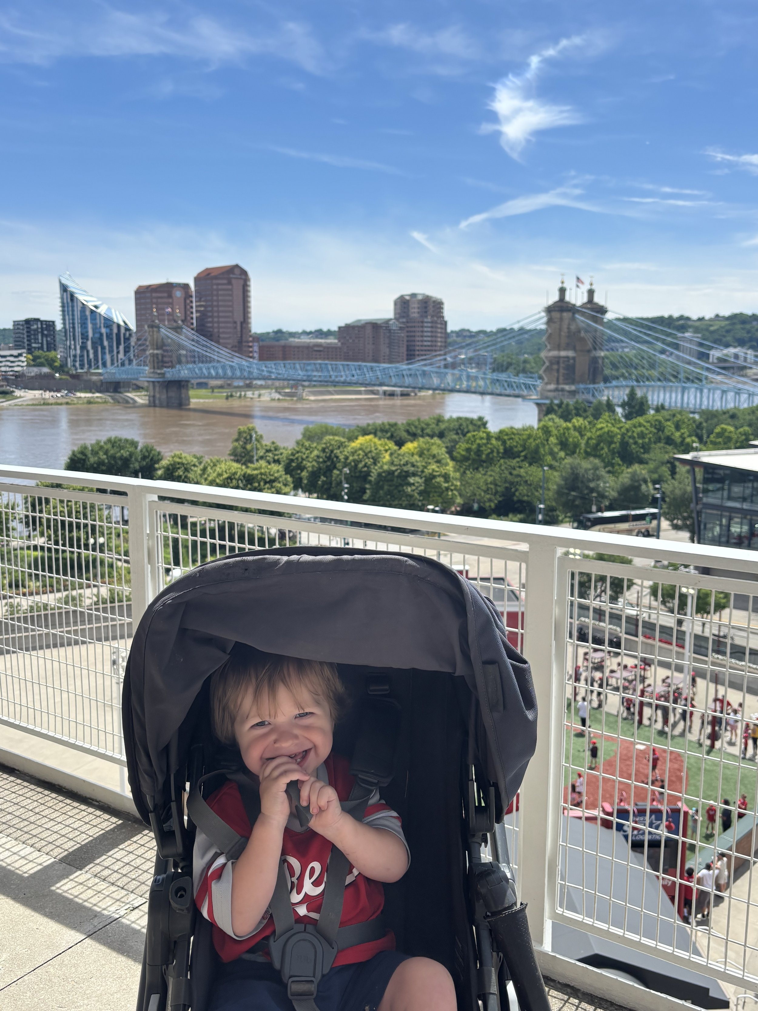 A young boy with light hair smiling and sitting in a stroller on an outdoor balcony, with a view of a river, a bridge, and city buildings under a partly cloudy blue sky.