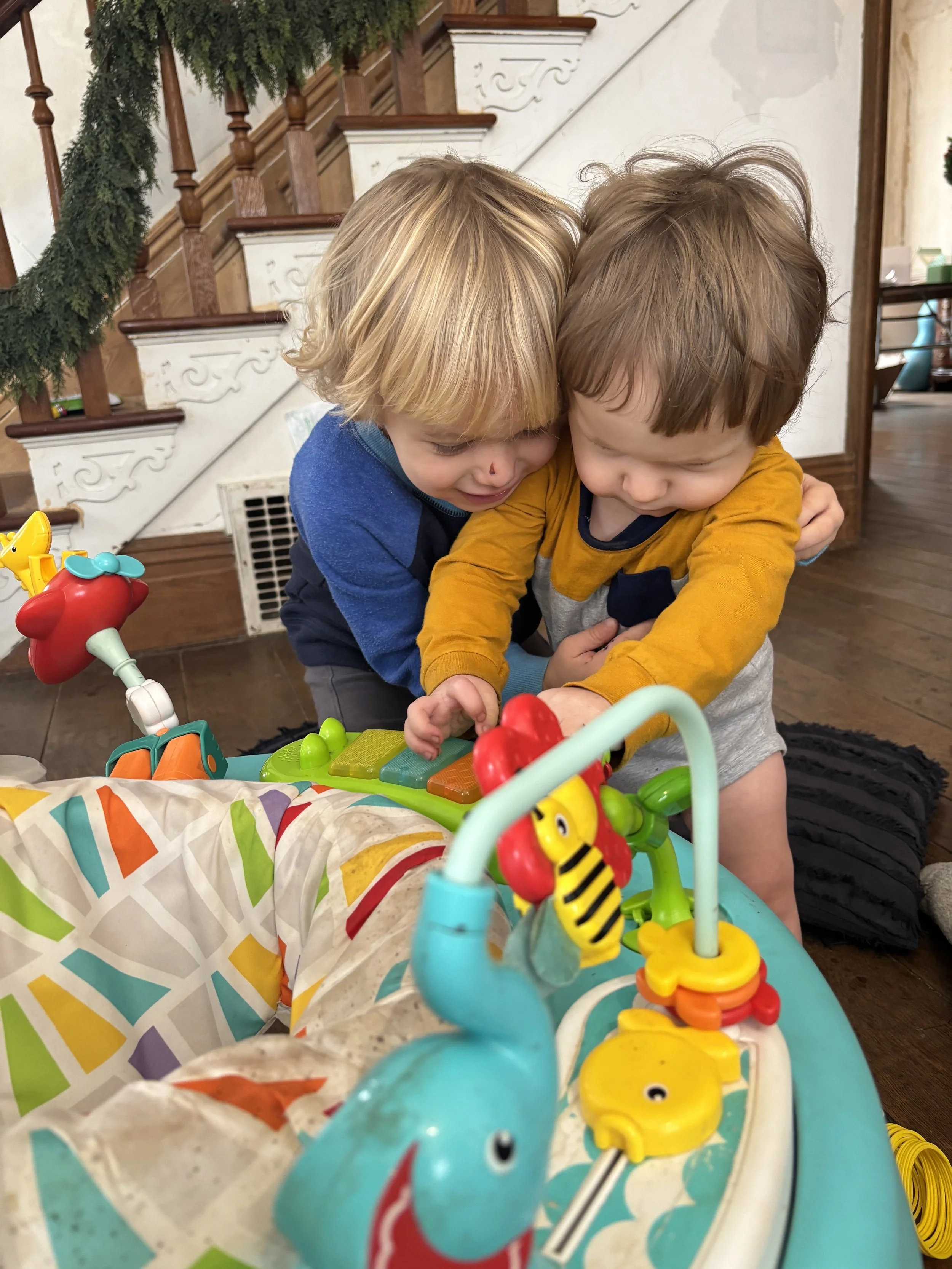 Two young children hugging and playing together in a cozy indoor setting near a staircase with festive greenery. They are engaging with a colorful, toy activity center featuring animals and various shapes.