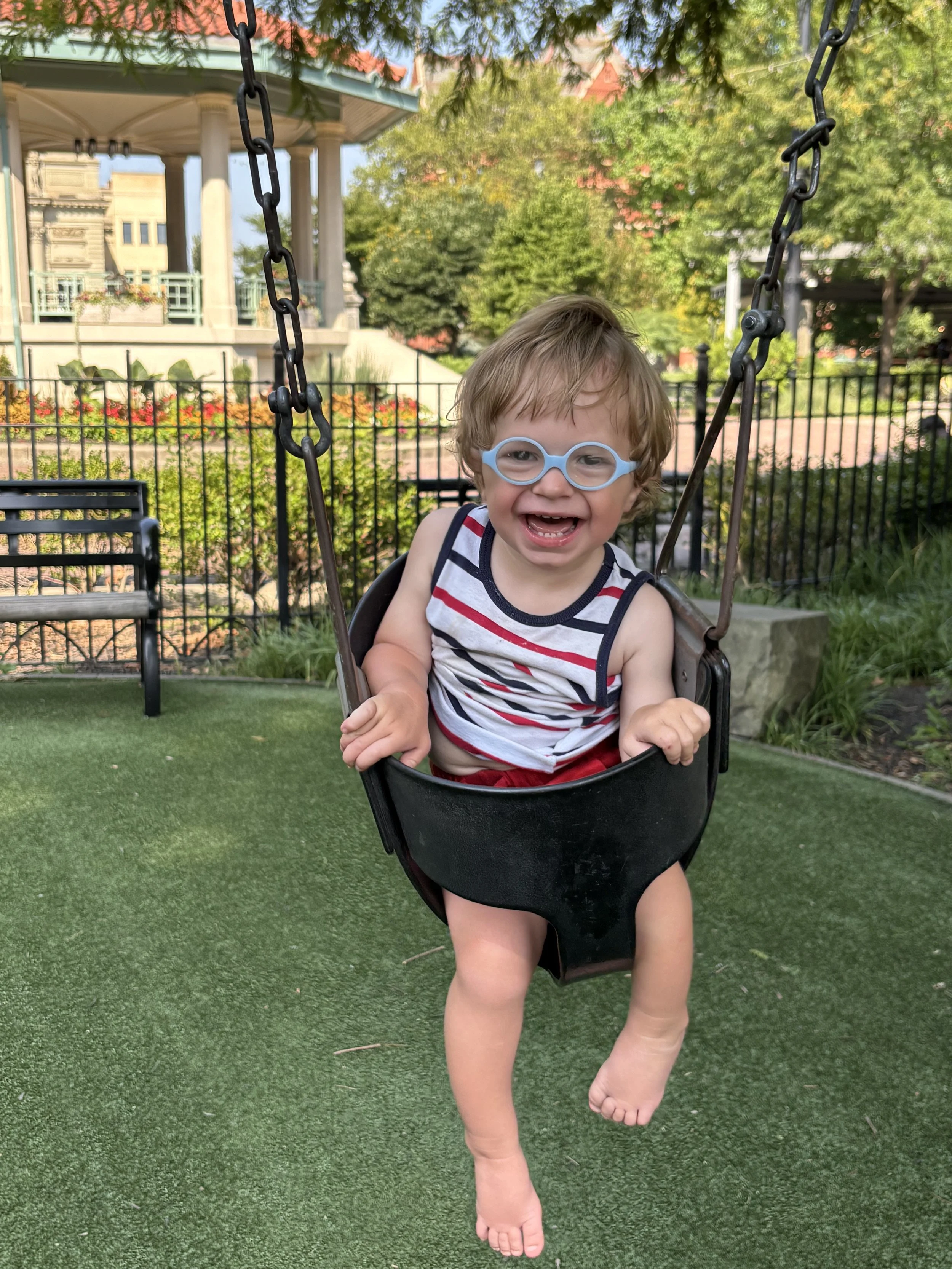 A young boy with glasses and a striped tank top smiling and swinging on a playground swing on a sunny day with trees and buildings in the background.