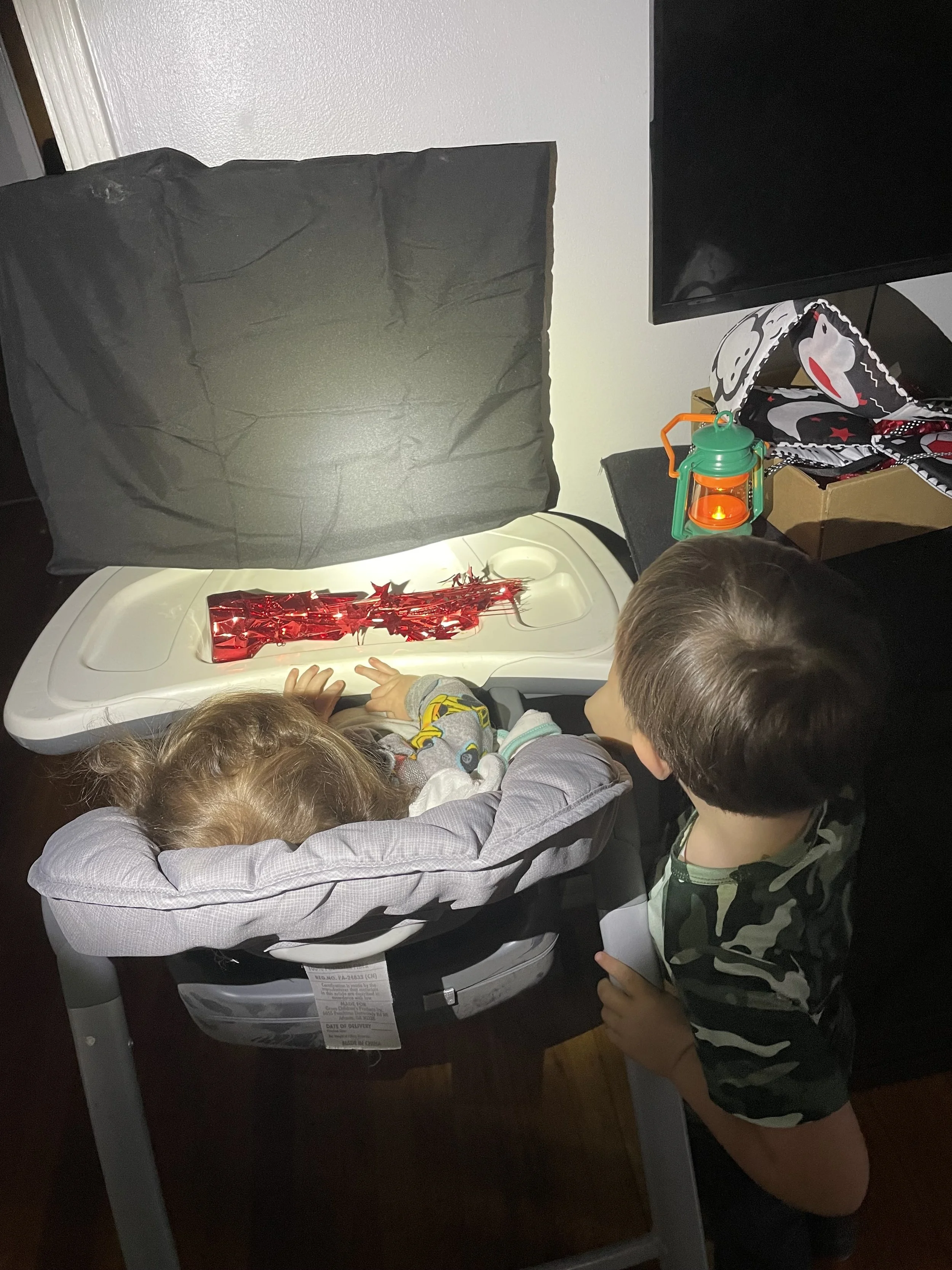 A baby sits in a high chair facing the wall. The wall has a black sheet on it. On the tray is a shiny red table decoration. Another boy stands next to high chair with toy lantern. 