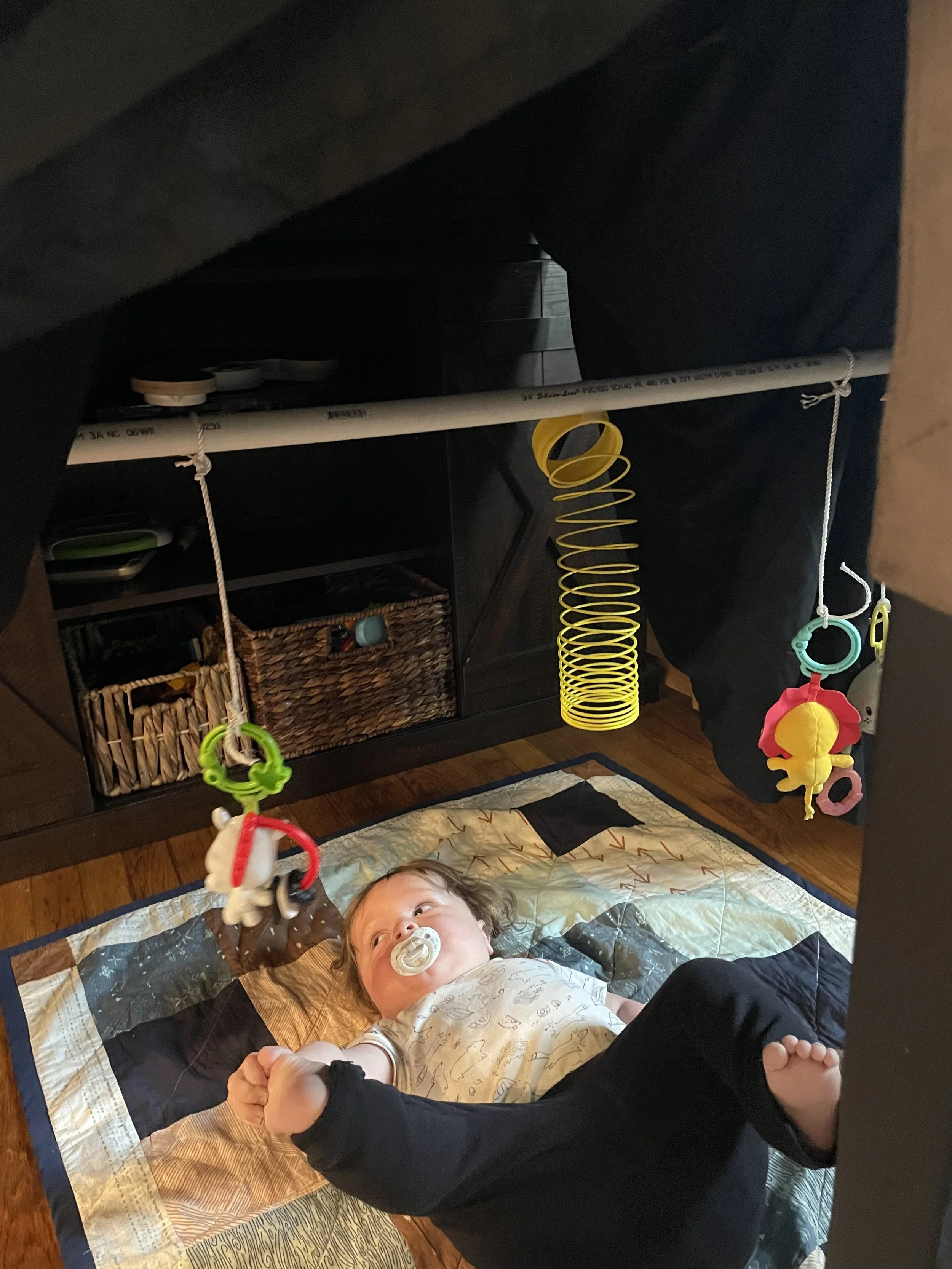 Baby lays on square pattern quilt. Above him hangs various baby toys. 