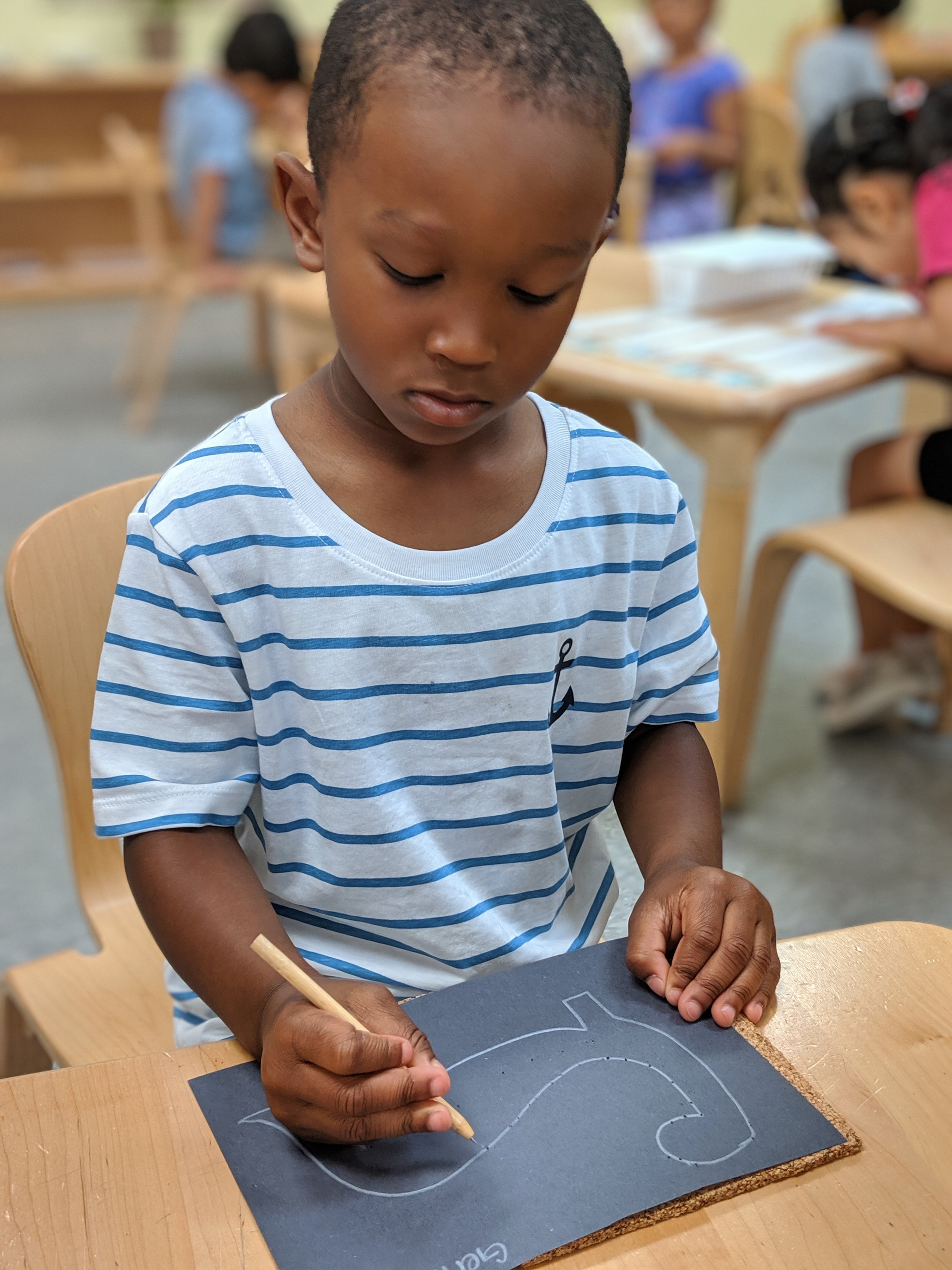 A young boy sitting at a table in a classroom, drawing with a wooden stick on black paper. He is wearing a white and blue striped shirt with an anchor emblem. Other children are visible in the background, engaged in similar activities.