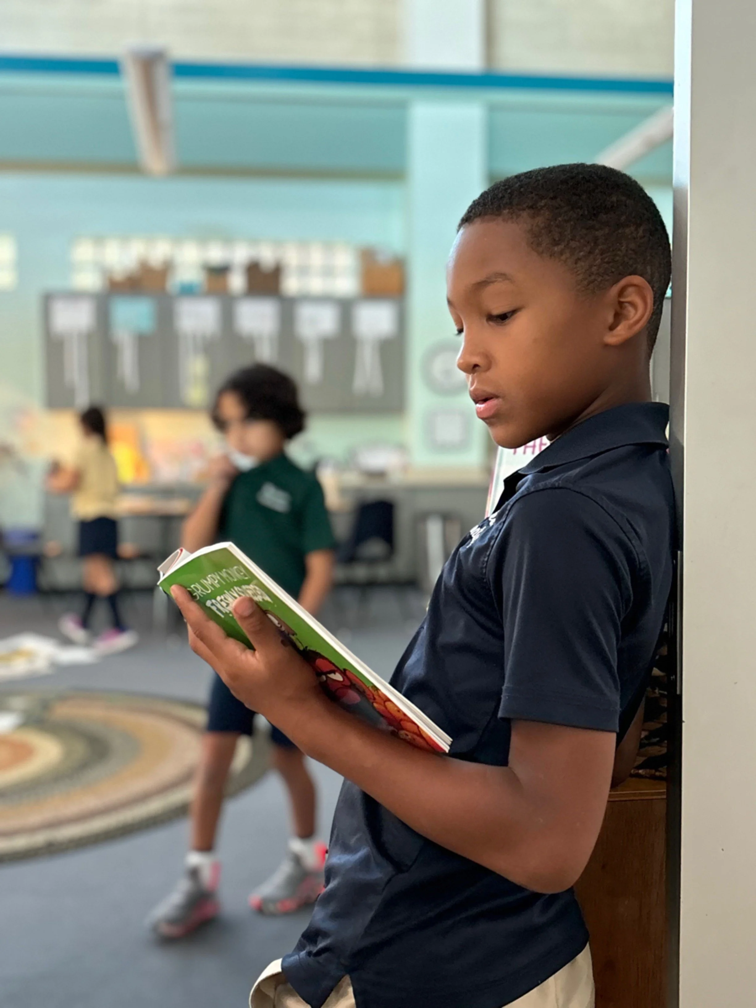 A student is focused reading a book while standing against a wall in a classroom.
