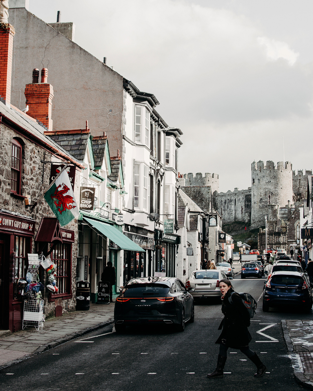 Street view of Conwy, Wales, with shops, traffic, a Welsh flag, and Conwy Castle in the background