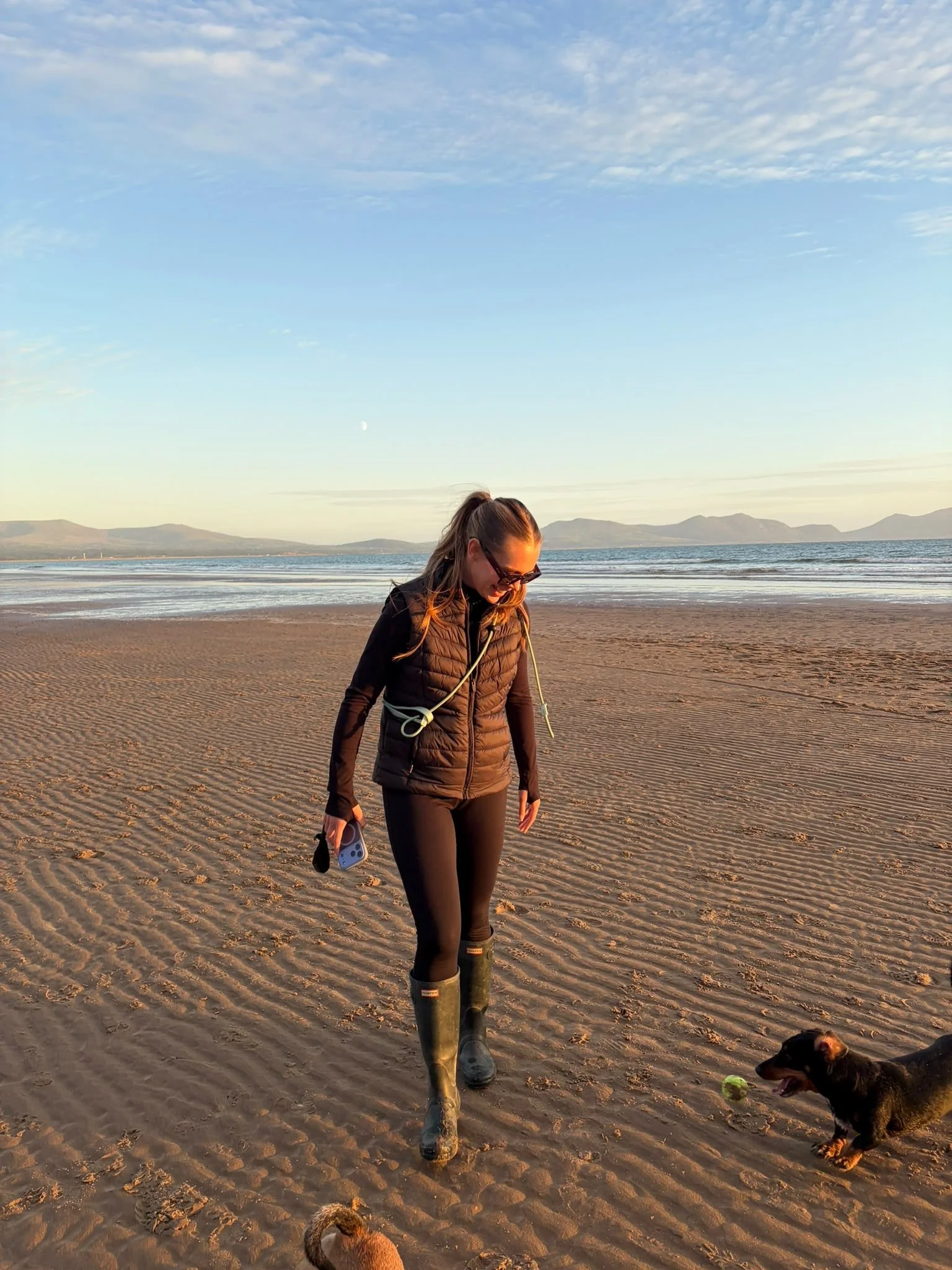 Woman in sunglasses and boots walking a dog on a sandy beach at sunset, with mountains and ocean in the background