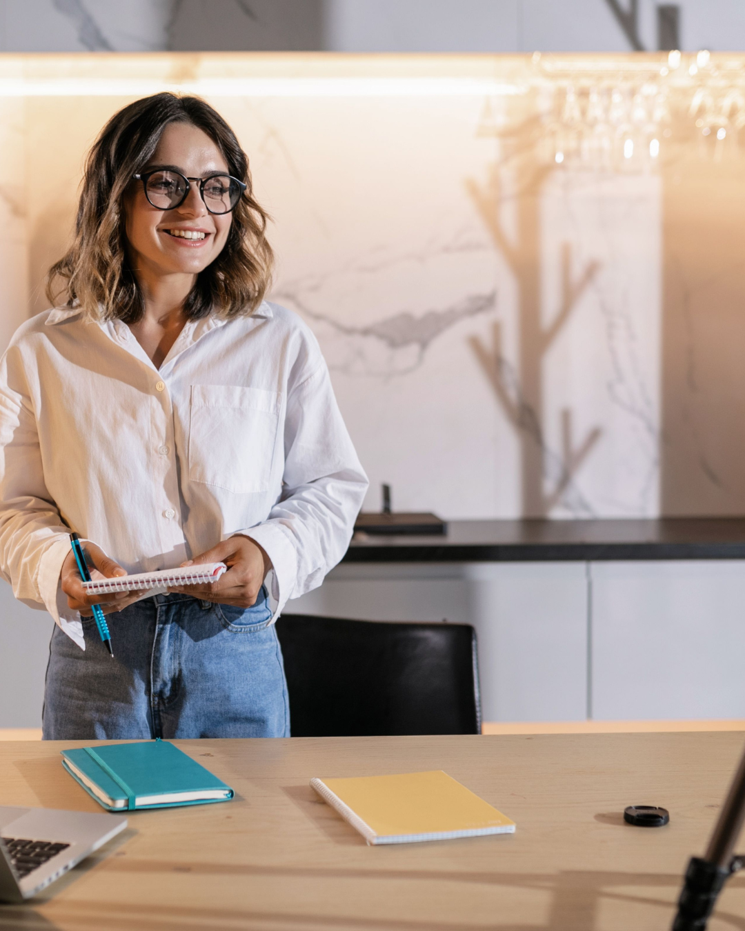 Smiling woman wearing glasses holds a notebook and pen in a modern office.