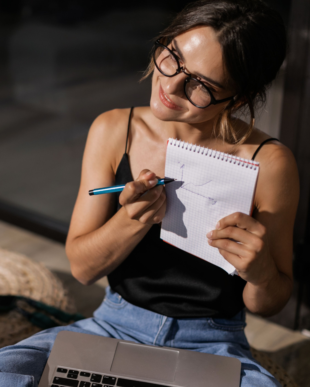 Woman wearing glasses writes a math graph in a spiral notebook beside a laptop