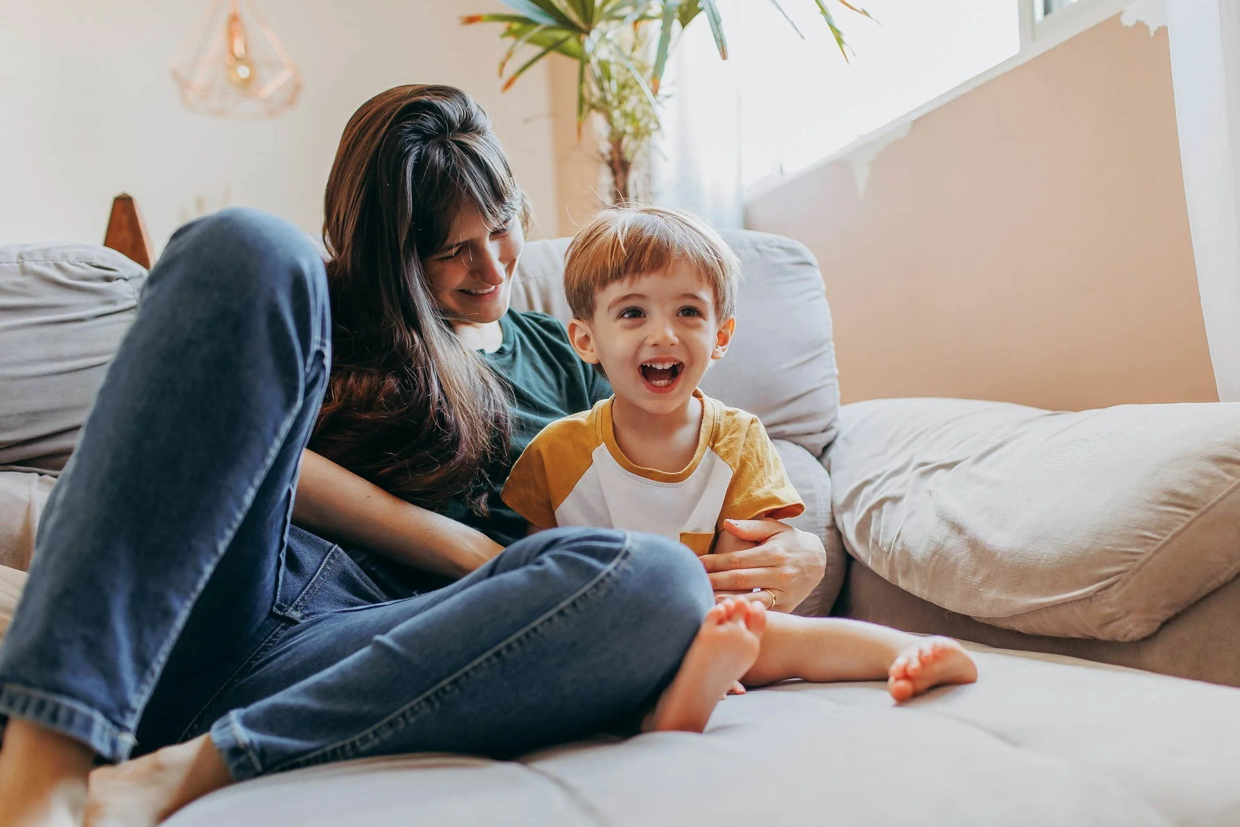A woman with long dark hair and a young boy with light brown hair are sitting on a light-colored couch in a brighter living room. The woman is smiling and leaning toward the boy, who is laughing with his mouth open. There's a potted plant near the window and a geometric pendant hanging from the ceiling.