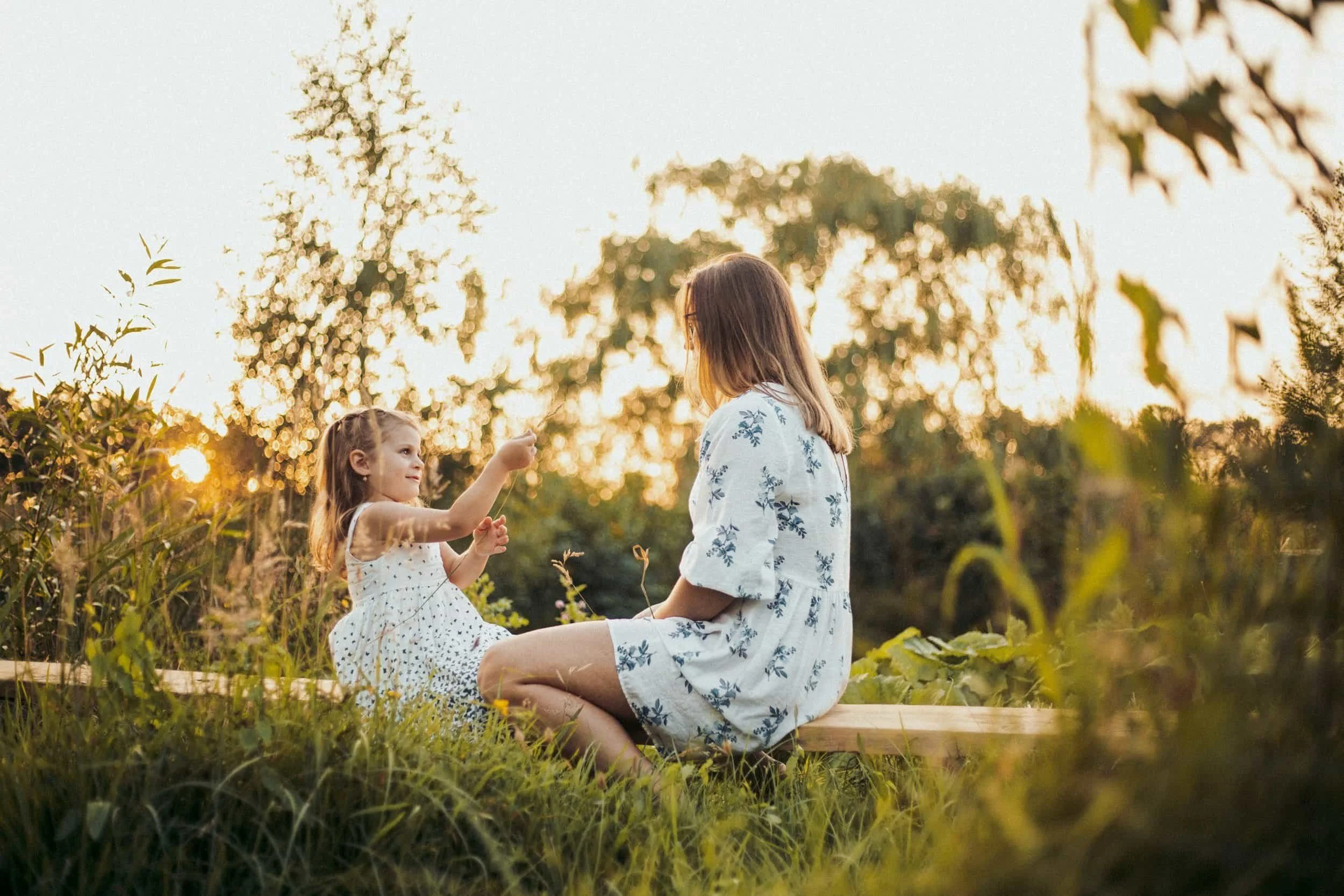 A woman and a young girl, possibly mother and daughter, sitting on a wooden bench in a grassy field during sunset. The girl is handing a flower to the woman, and they are both smiling. The background features trees and a soft, glowing sky.
