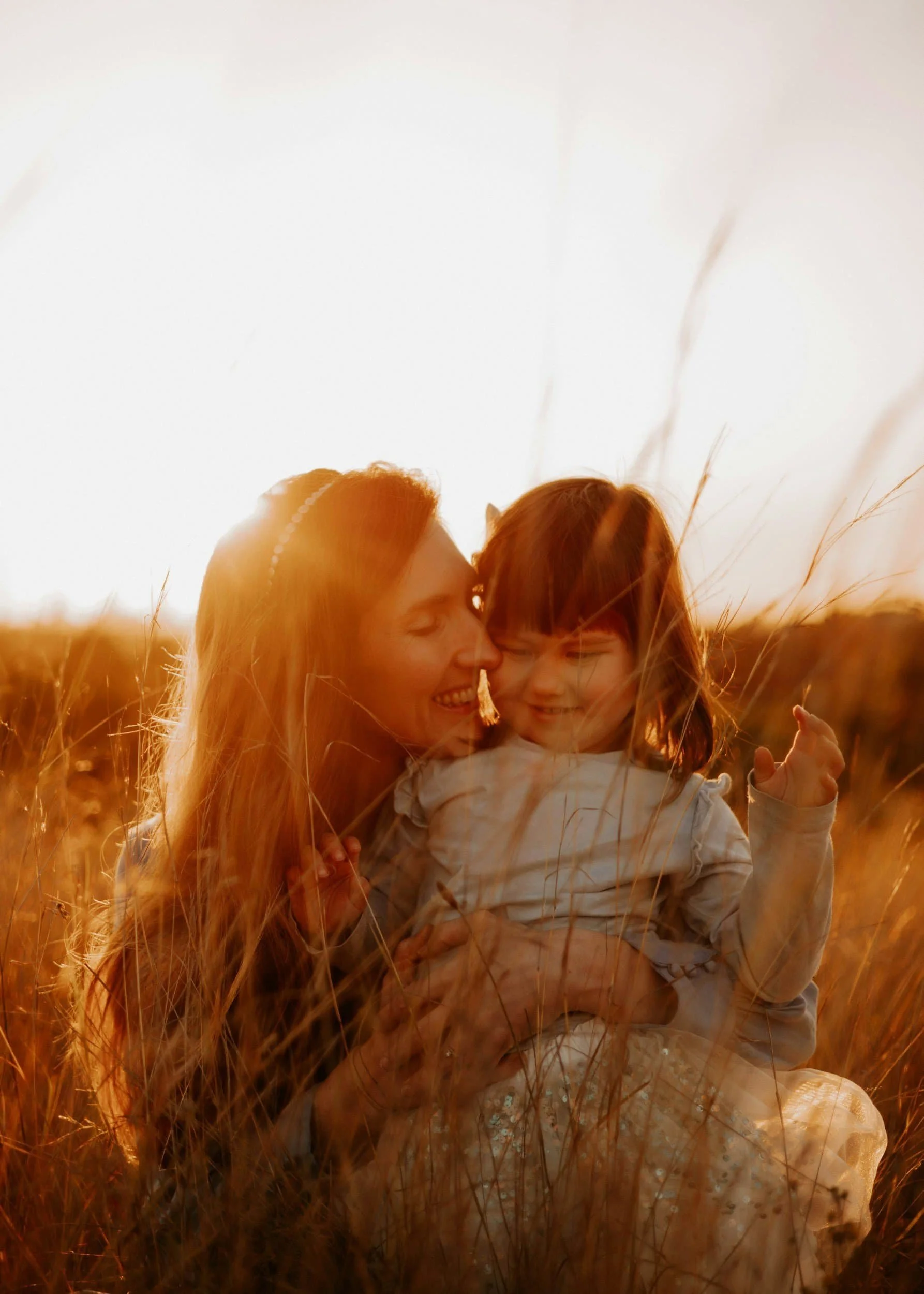 A woman and a young girl sharing a close moment in a wheat field at sunset, smiling and cuddling.