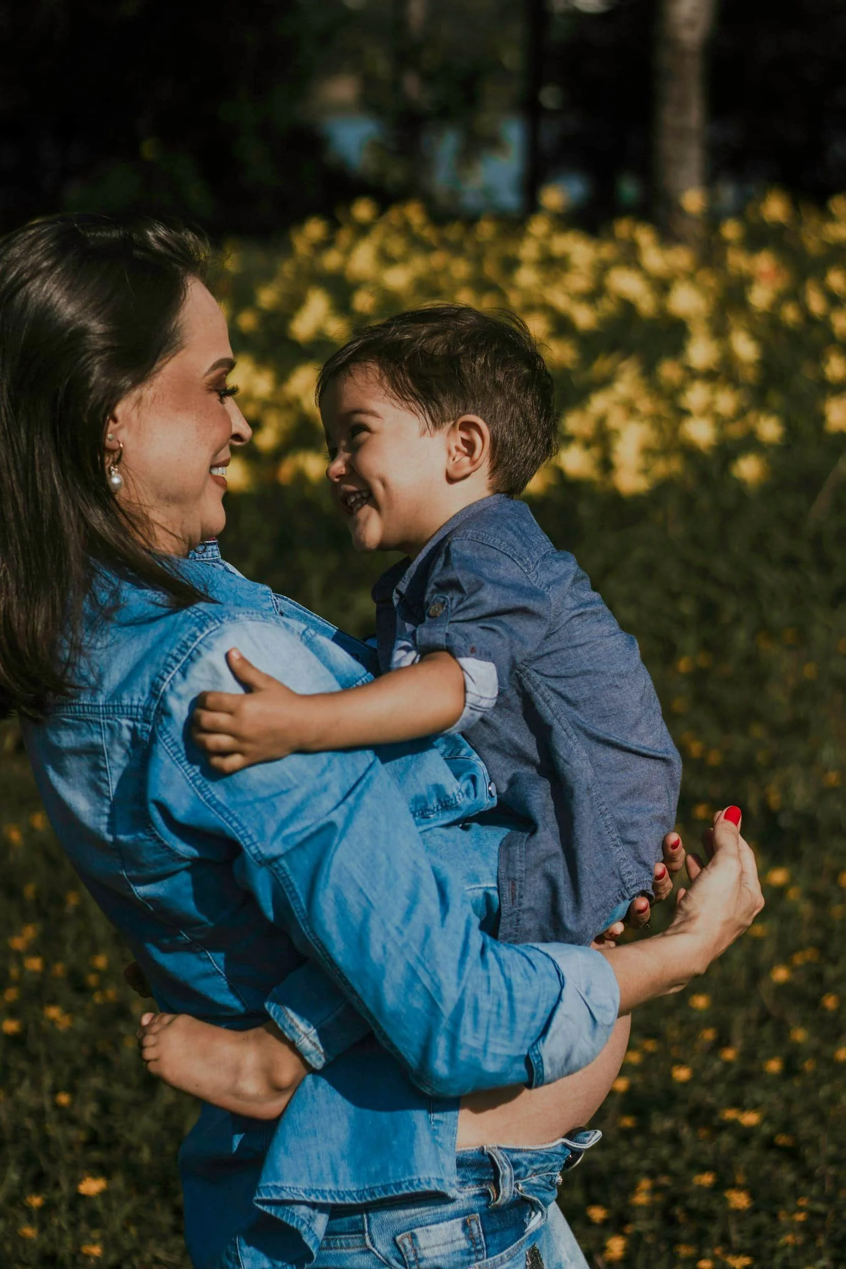 A woman with dark hair, wearing a denim jacket, holding a young boy with short dark hair and a denim shirt, as they smile and look at each other outdoors at sunset, with blurred trees and yellow flowers in the background.