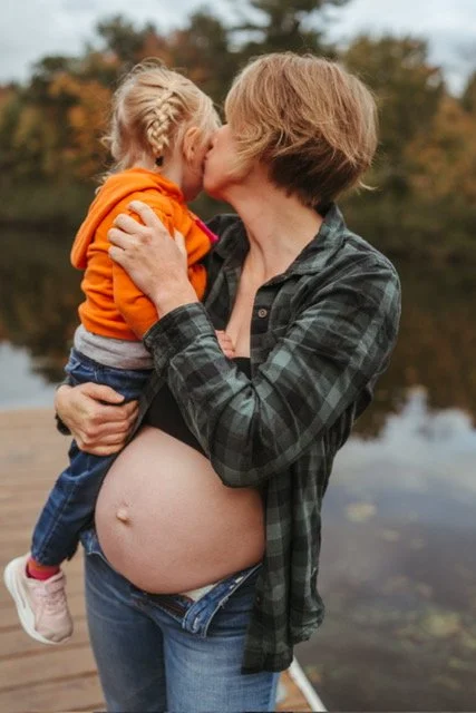 A pregnant woman holding a young girl, possibly her daughter, by a body of water during autumn. The woman is kissing the girl, who is wearing an orange hoodie.