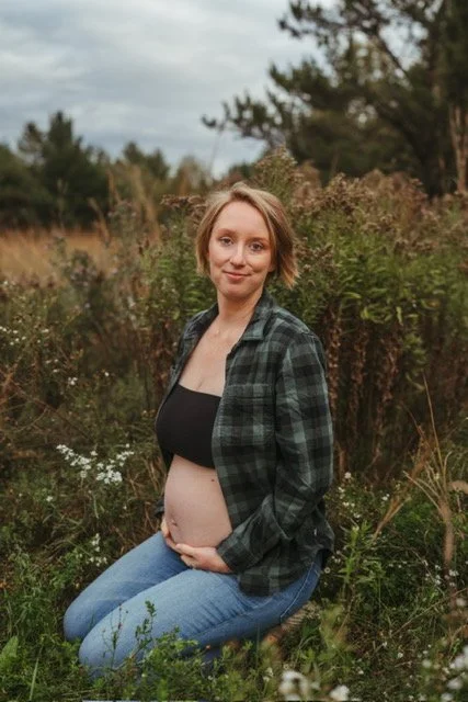 A pregnant woman with short blonde hair kneeling outdoors in a grassy field with bushes and trees, wearing a black top, a green plaid shirt, and blue jeans.