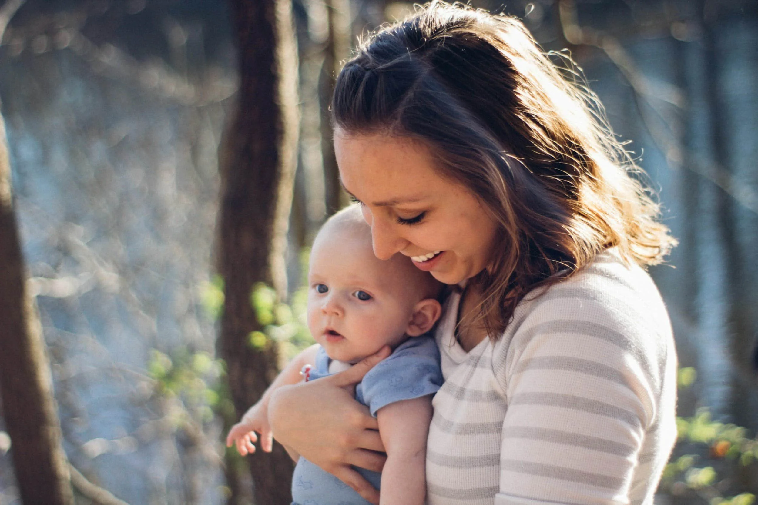 A woman with brown hair smiling while holding a baby outside among trees.