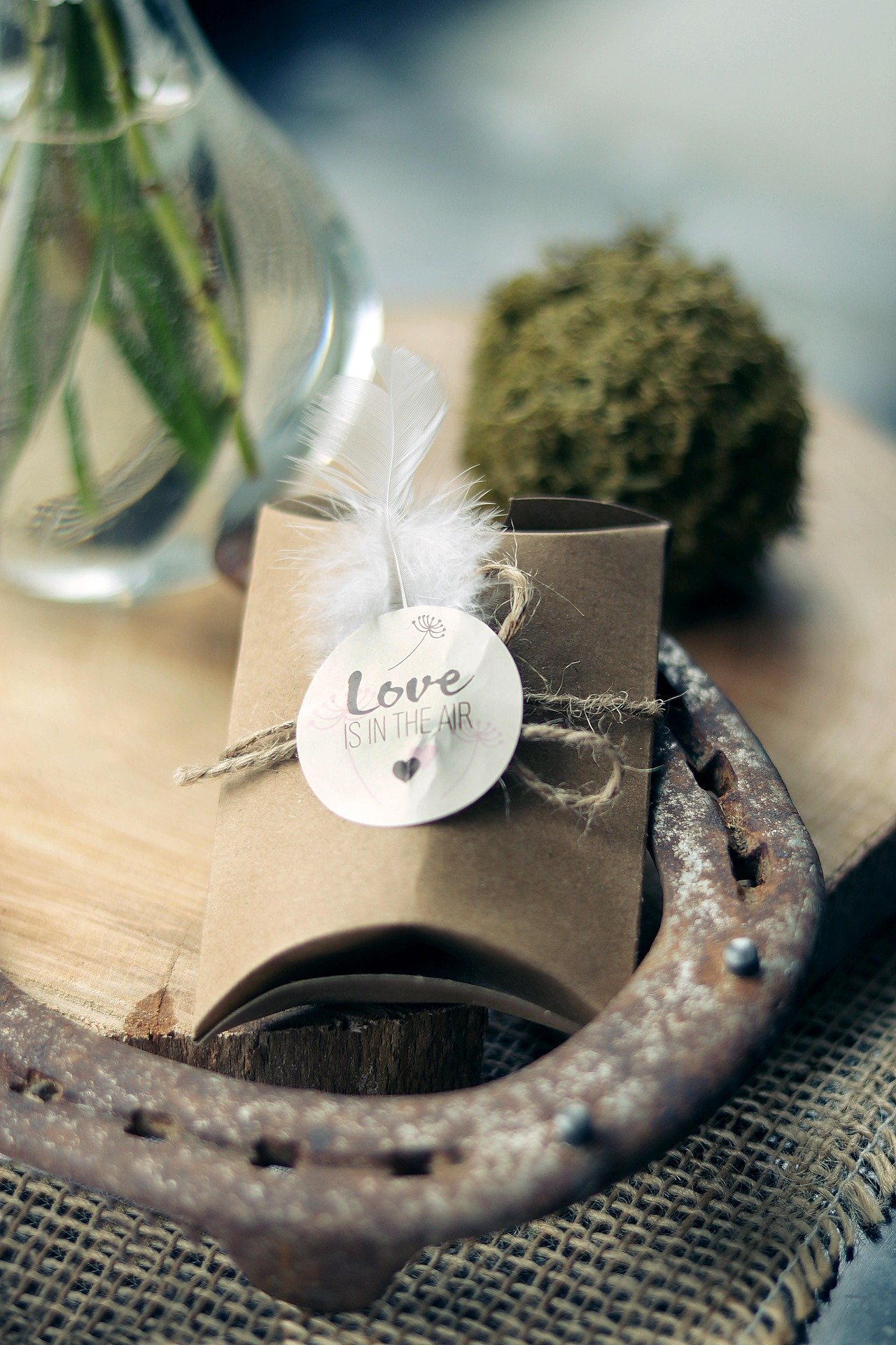 A small gift wrapped in brown paper with a white feather and a round tag that says "Love Is in the Air," placed inside a rusty metallic horseshoe on a wooden surface.