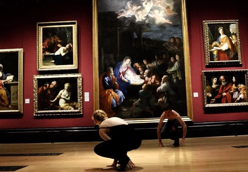 Two people crouching in front of large religious paintings in an art gallery with red walls and wooden flooring.