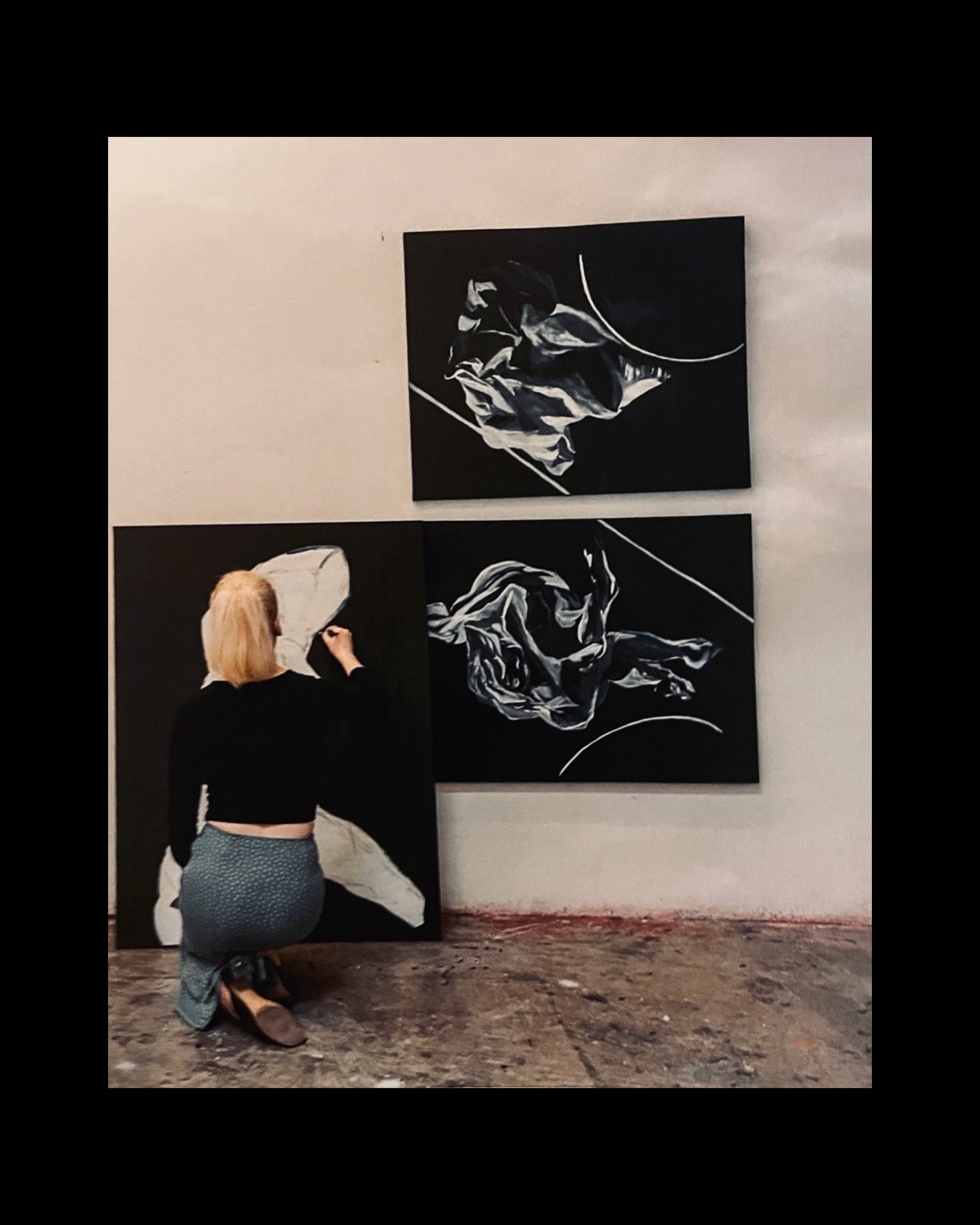 An artist kneels on a dusty floor, working on a large abstract black and white painting displayed on a white gallery wall. The artist has blonde hair, wears glasses, a black top, and patterned pants.