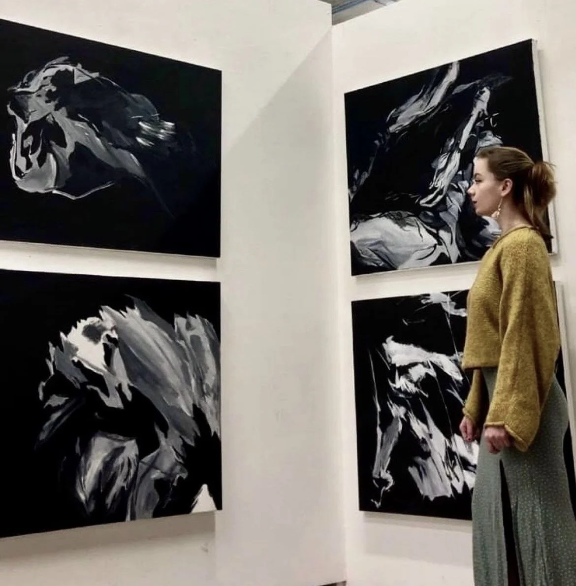A woman in a yellow sweater and gray skirt looking at black and white paintings of lions in an art gallery.