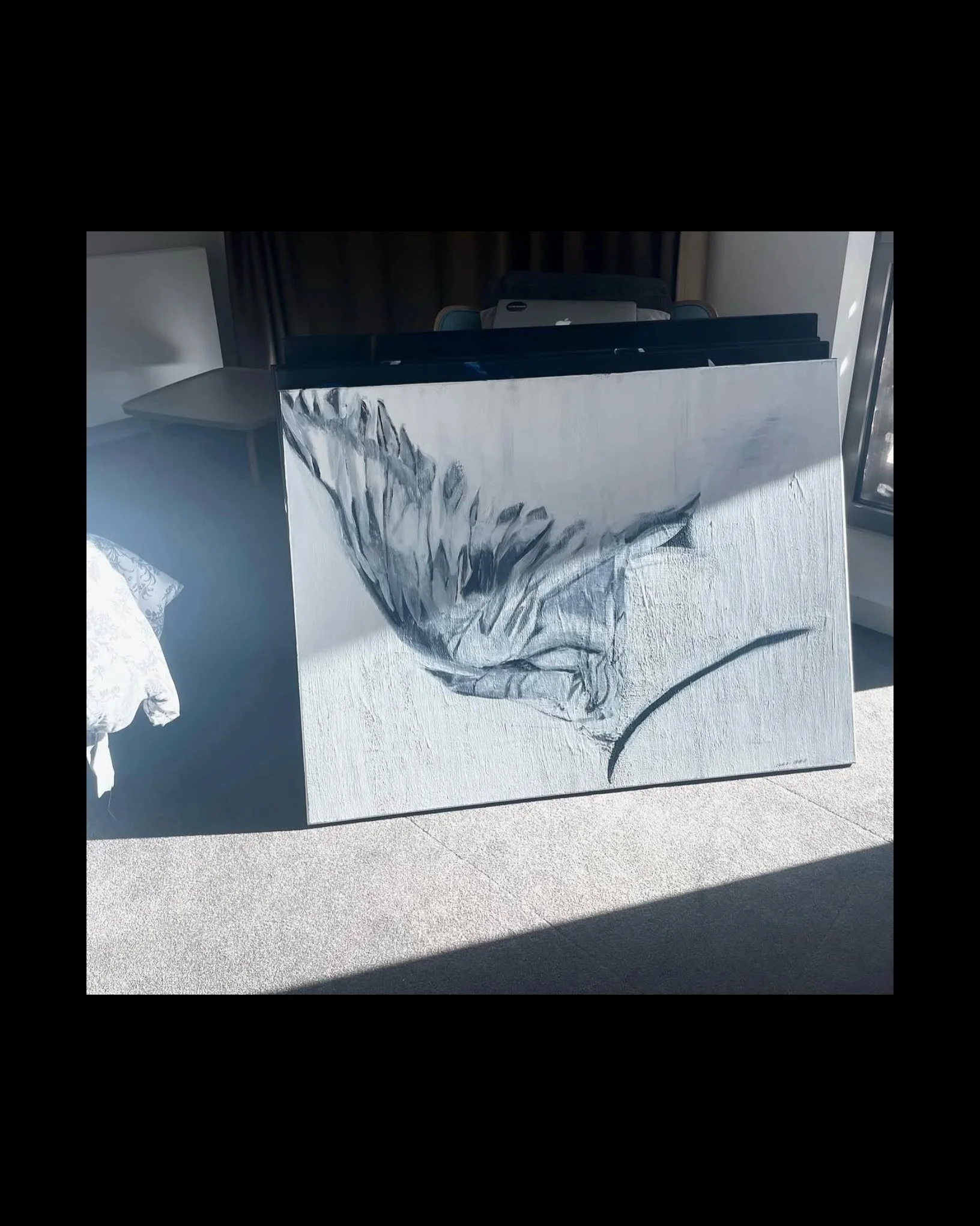 A canvas painting of a horse's head with a black and white color scheme is propped up against a surface. In the background, there's a laptop on a chair and a dark curtain, with sunlight casting shadows on the floor.