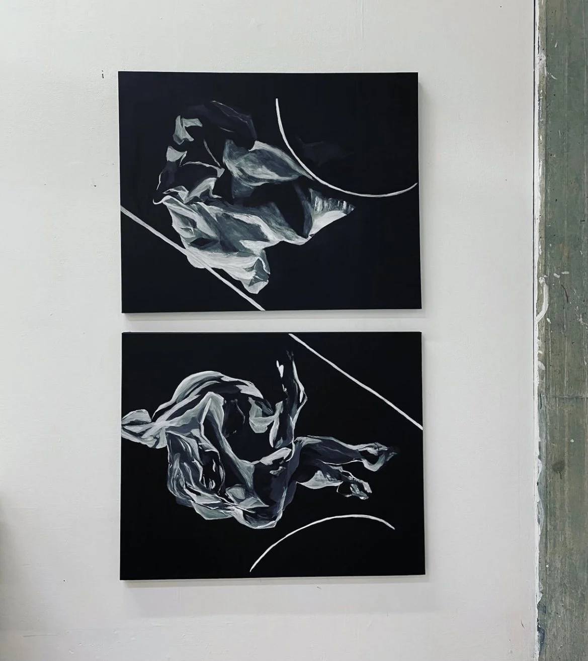 Two abstract paintings on a white gallery wall, featuring black backgrounds with swirling white and gray shapes, connected by curved white lines.