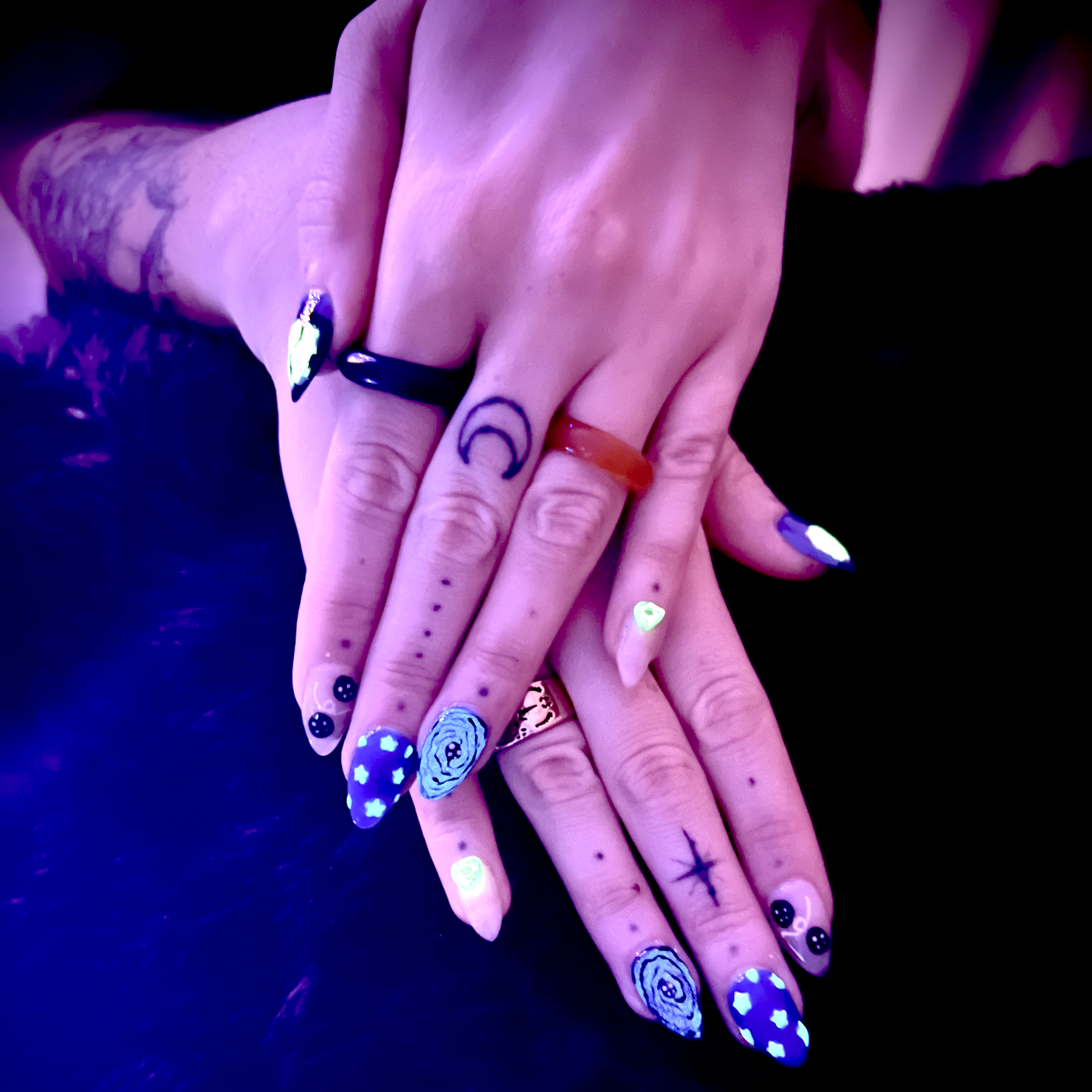 Close-up of a person with multiple fingers decorated with brightly colored, possibly glow-in-the-dark nail polish, tattoos, and jewelry, under a purple and blue lighting effect.