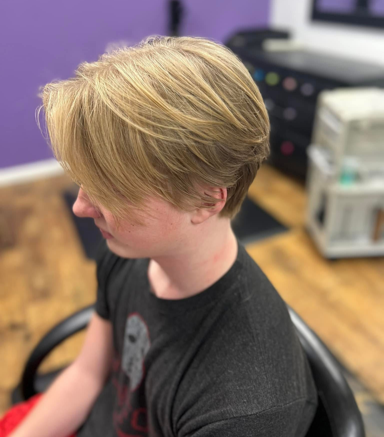 A person with short, layered blonde hair sitting in a salon chair, viewed from the side, with a purple wall and salon equipment in the background.