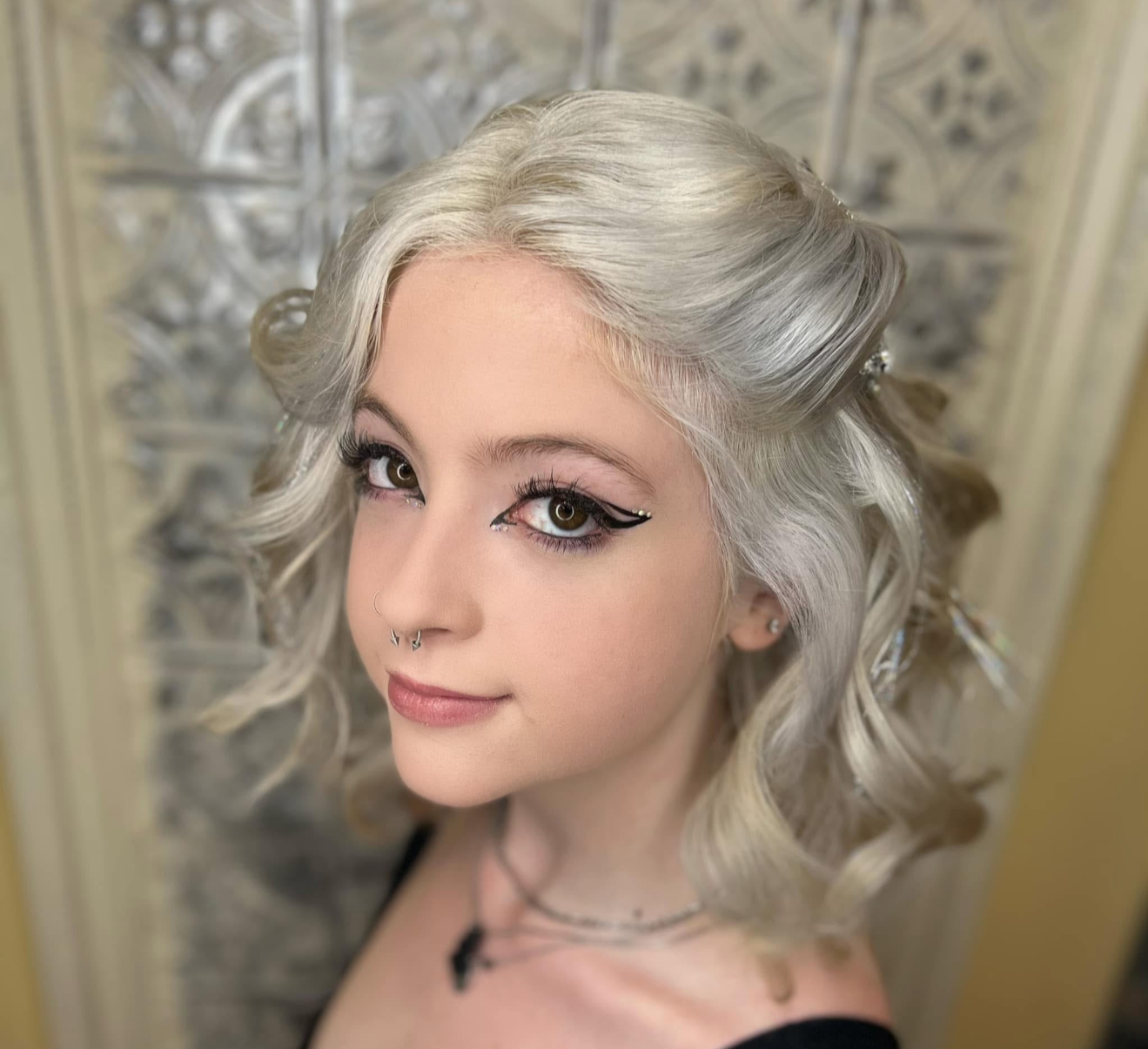 A young woman with platinum blonde hair styled in loose curls, wearing makeup with winged eyeliner and contact lenses, and jewelry including a nose ring and layered necklaces. She is looking at the camera with a slight smile, standing in front of a d