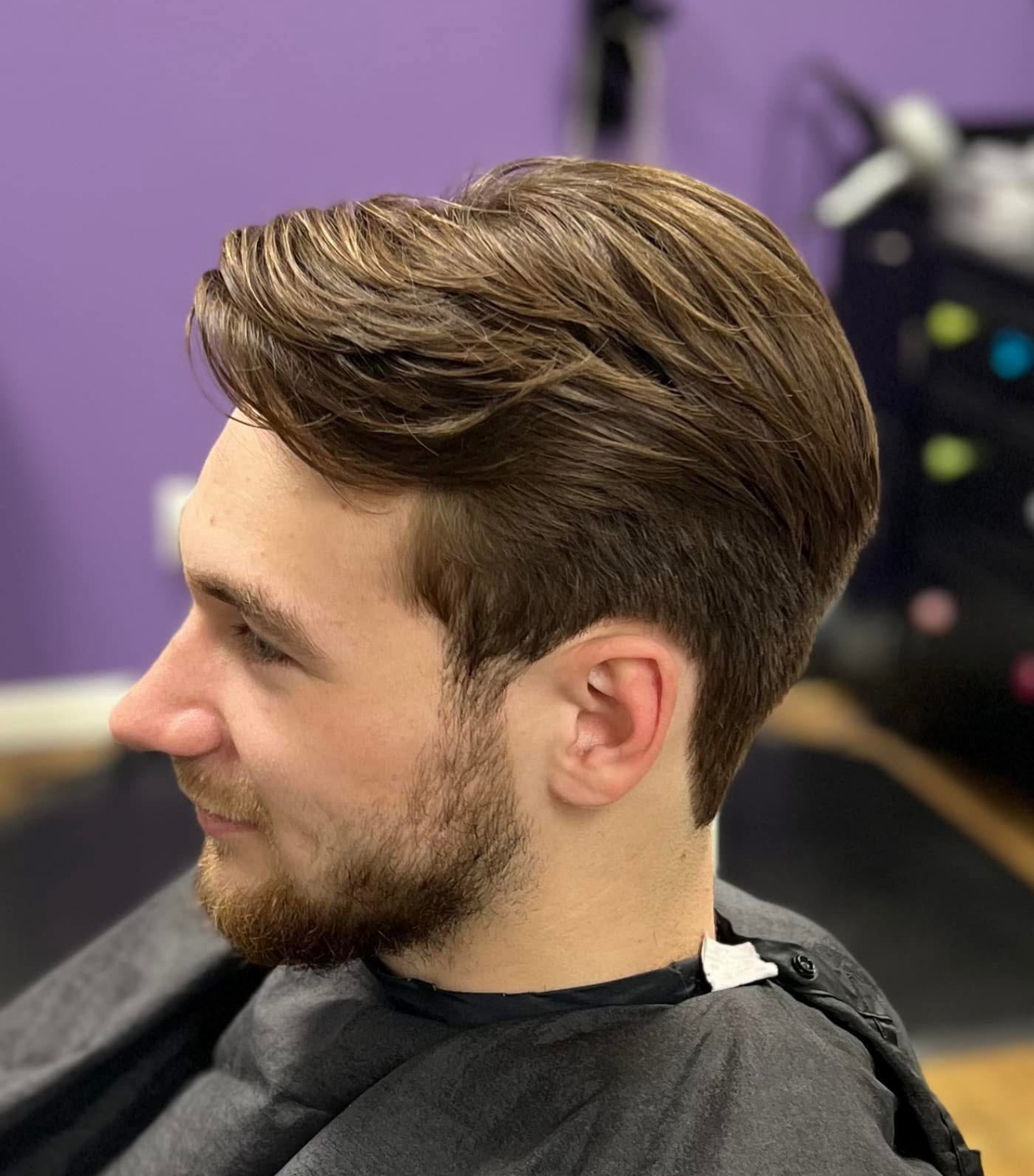 Side view of a young man with styled brown hair and a beard, sitting in a salon chair with a purple wall in the background.