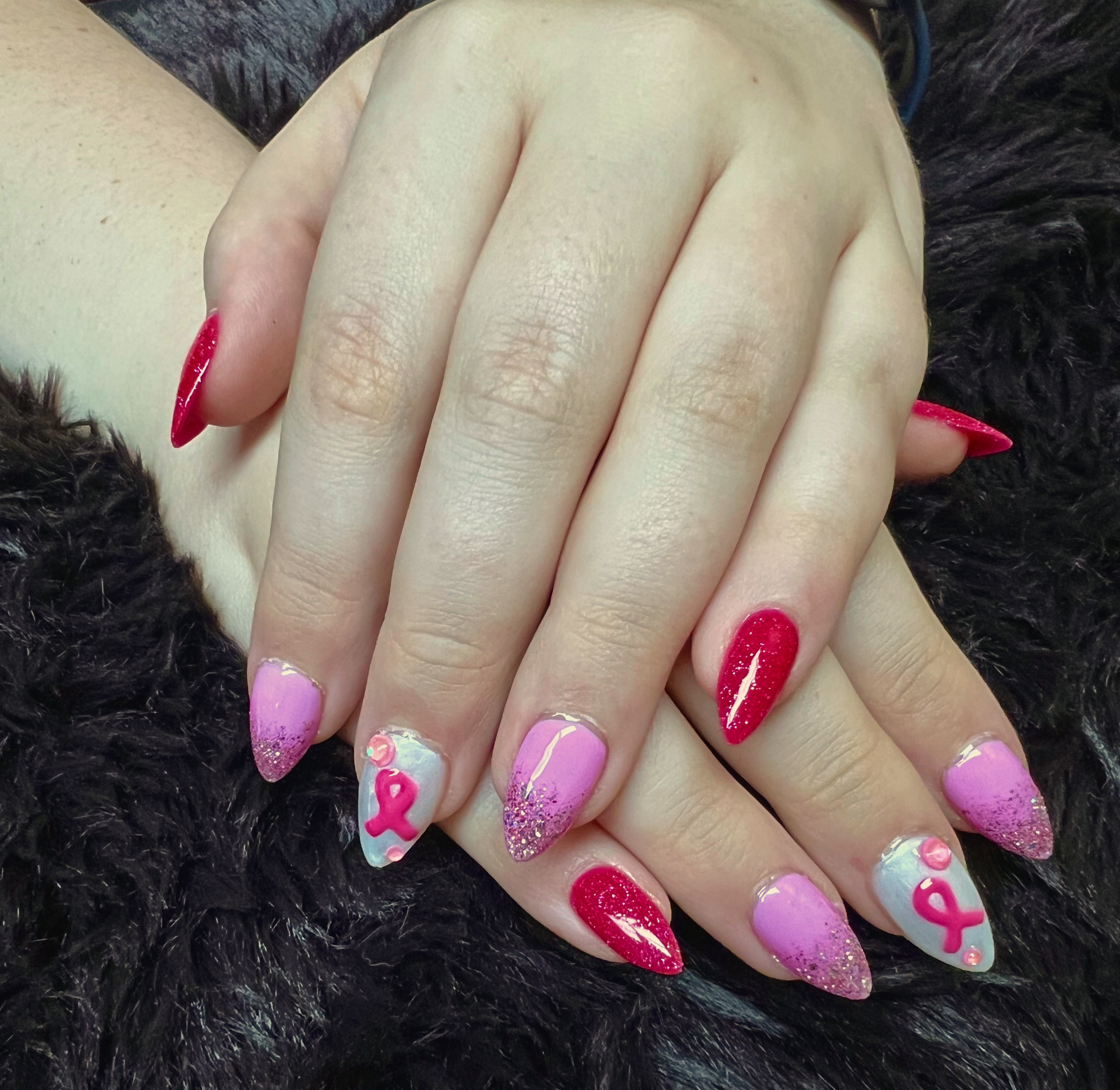 Close-up of a hand with decorated fingernails, with the index and ring fingers painted red and the middle, pinky, and thumb painted purple with glitter and heart designs. The person is wearing a black furry jacket or coat.