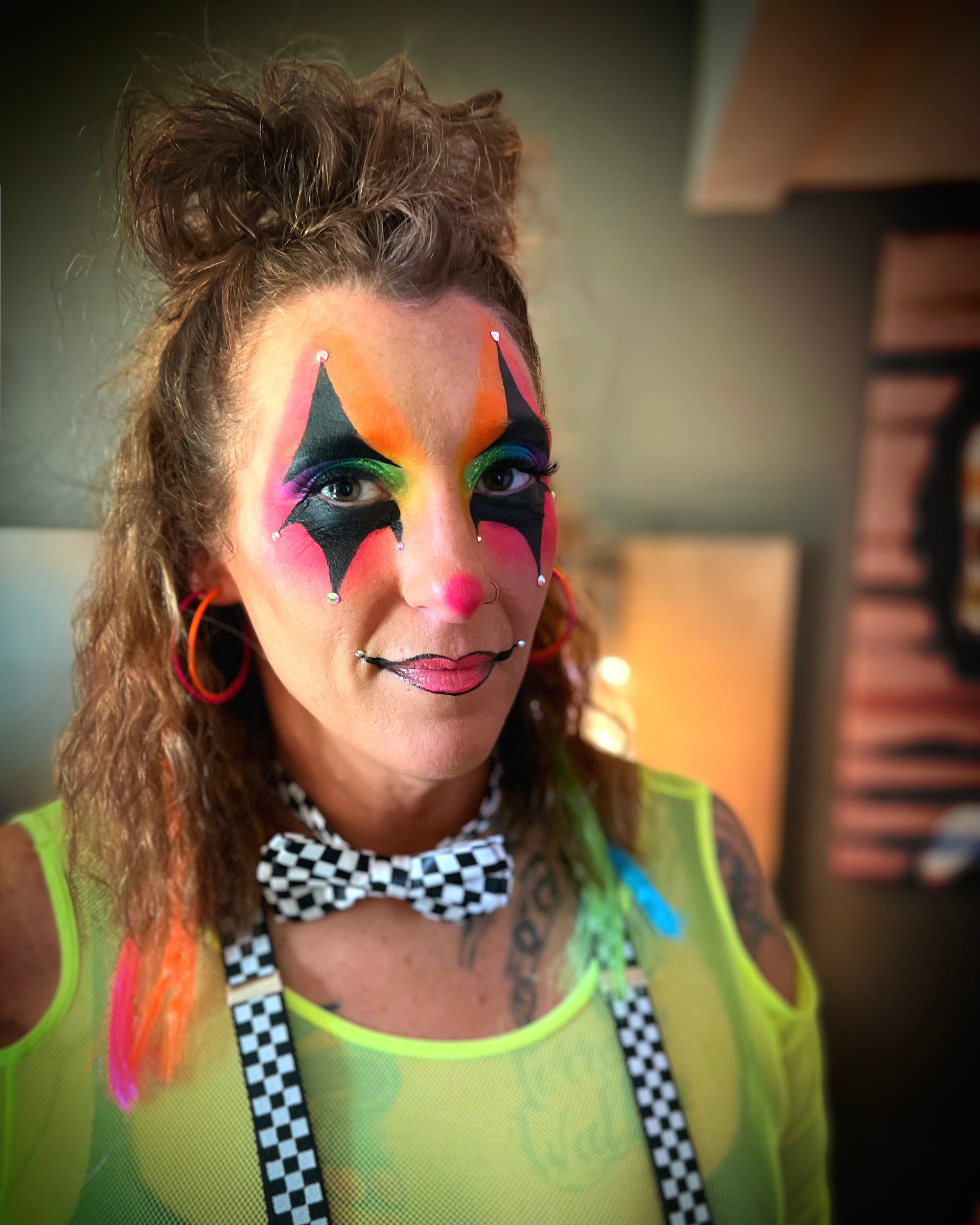 A woman with colorful clown makeup, including jagged black and pink designs around her eyes, a painted red nose, and pink lips. She has curly brown hair styled in a high bun and is wearing a neon green vest with checkerboard suspenders and a black an