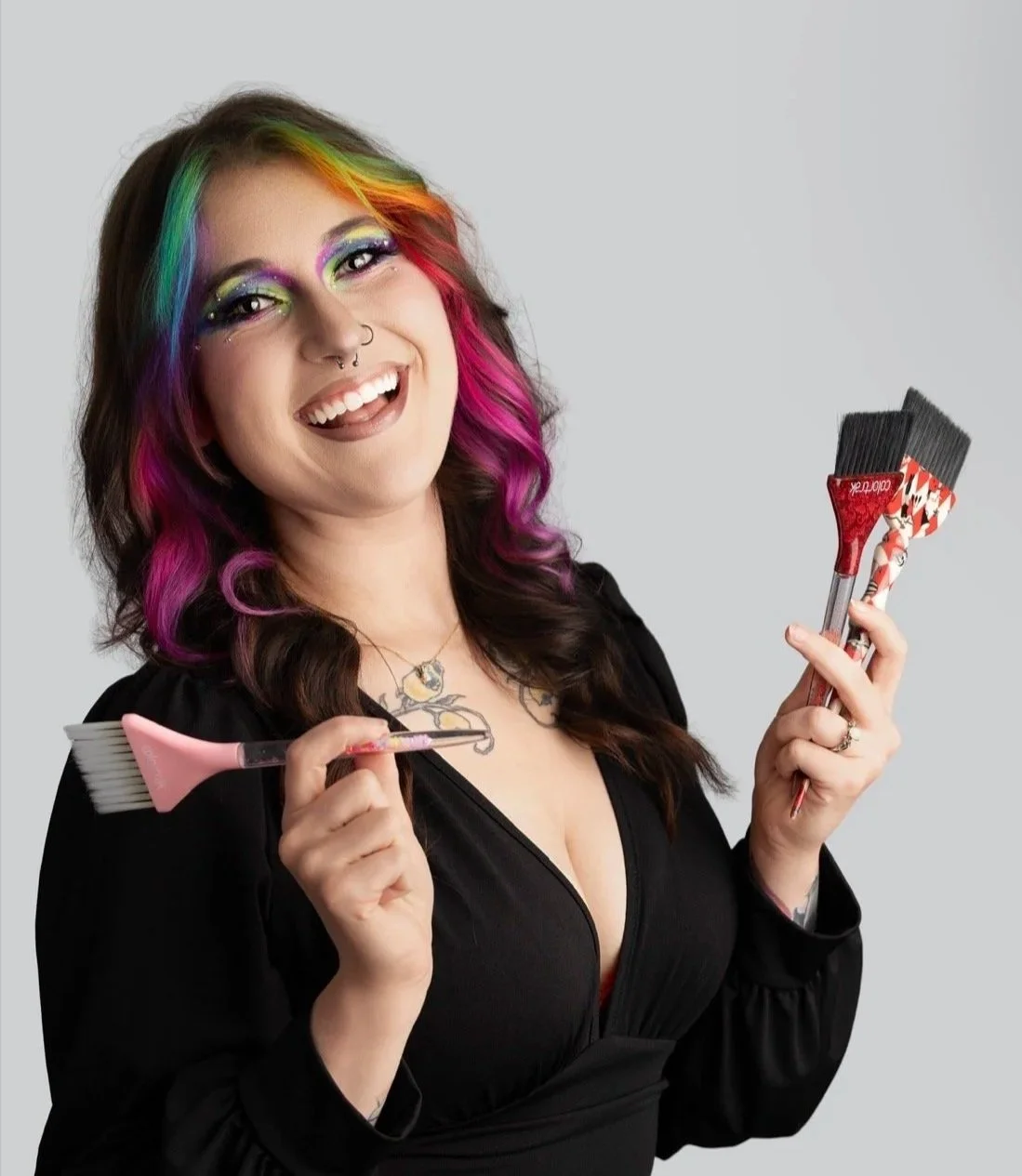 A woman with colorful rainbow makeup and dyed pink and purple hair holding a small paintbrush and two hair dyeing brushes, smiling against a plain gray background.