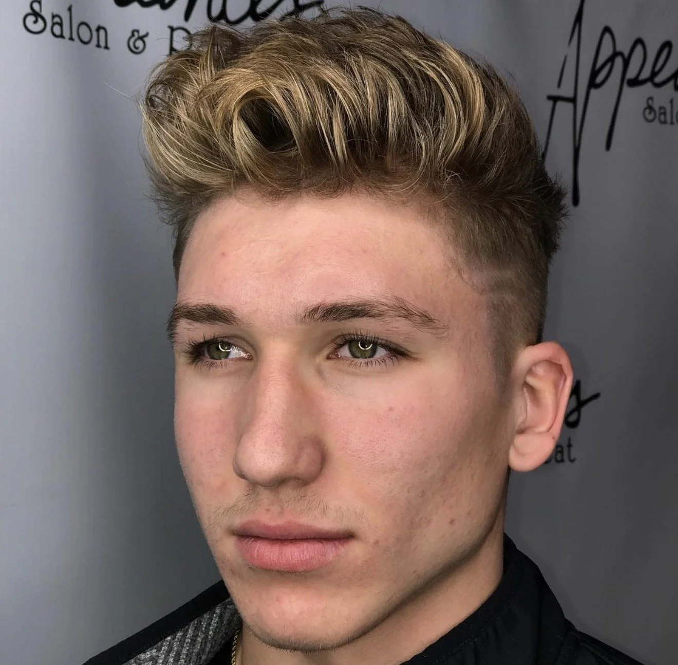 A young man with styled blonde hair and green eyes looks to the side against a gray background featuring salon branding.