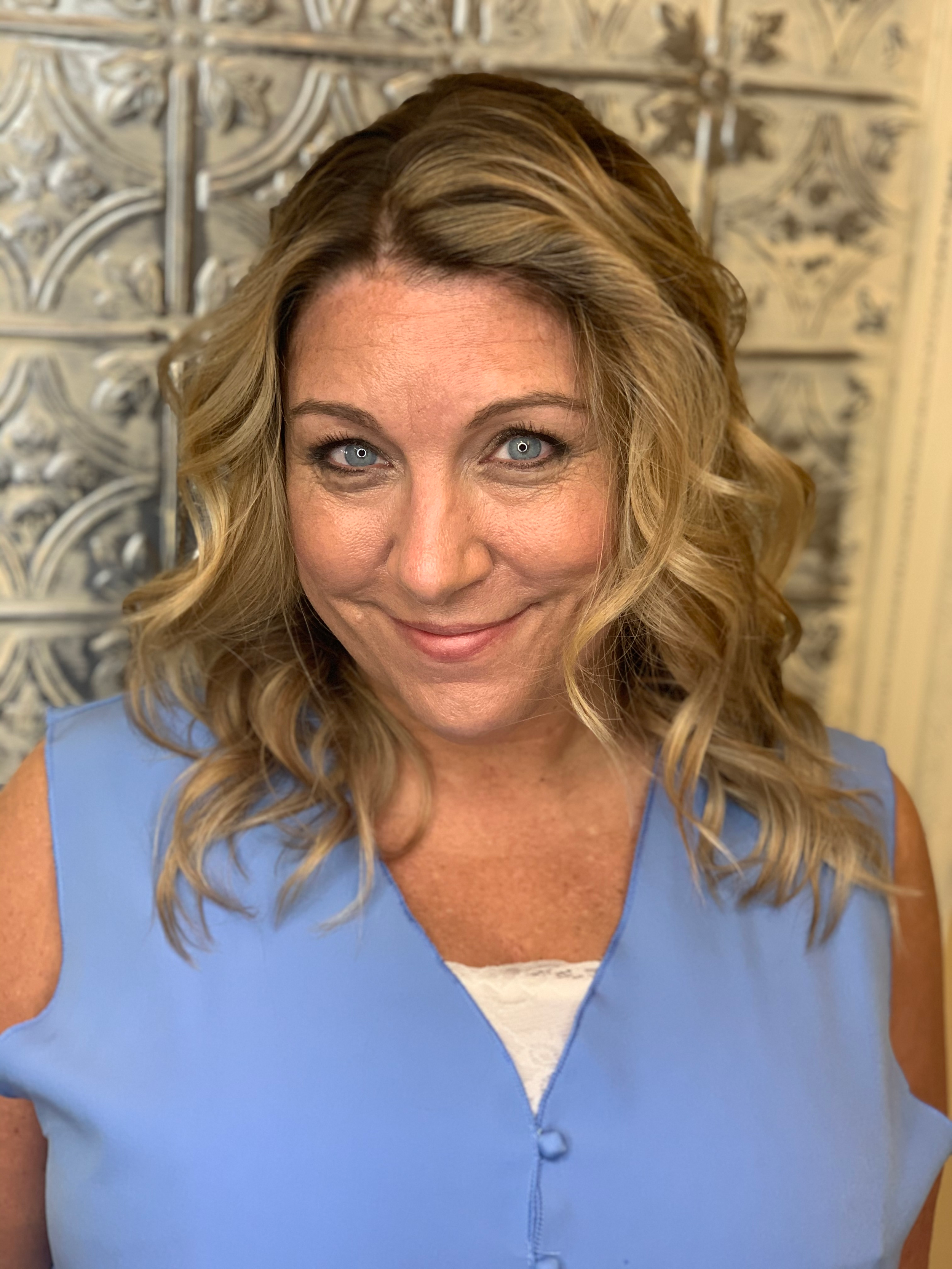 A woman with curly blonde hair styled in loose waves, wearing a light blue sleeveless top with a white shirt underneath, standing in front of an ornate, decorative silver wall.