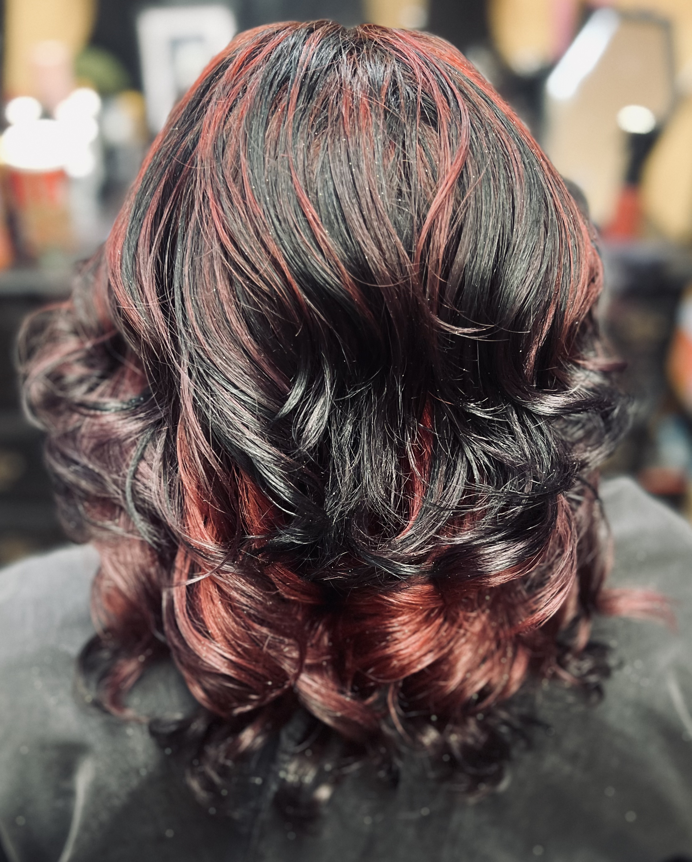 Close-up of a mannequin head with styled and colored hair featuring black, red, and pink shades, with some highlights and curls, set against a blurred background.