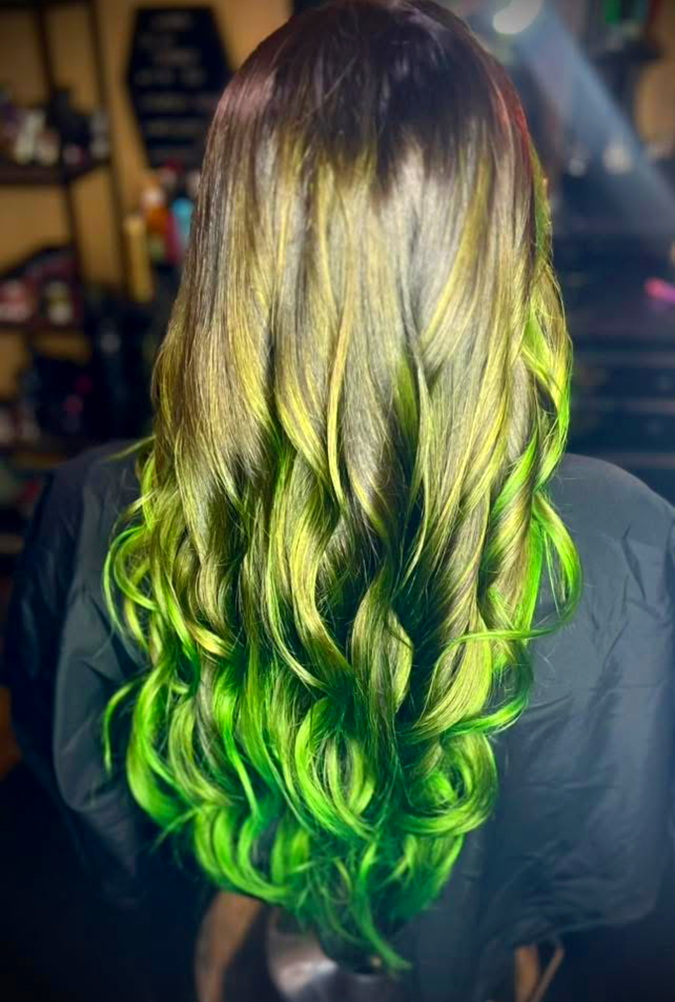 A person with long, wavy hair featuring glowing green and yellow highlights, seen from the back in a dimly lit room.
