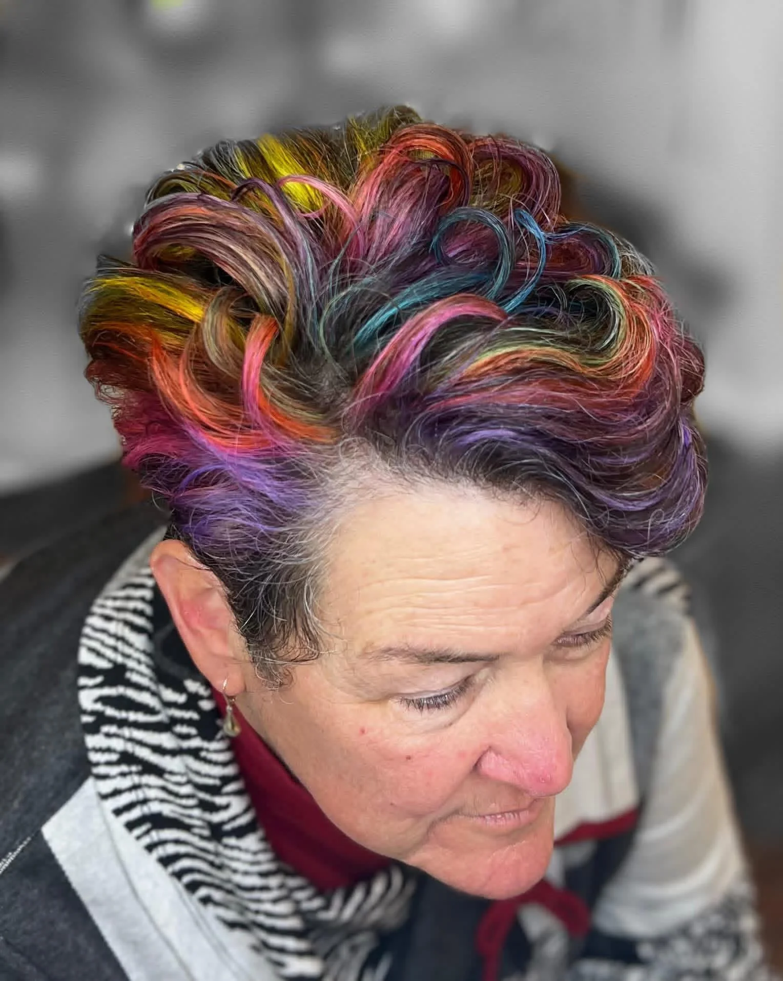Close-up of a person's head with colorful, rainbow-like hair styled in waves and curls, with a gray background.