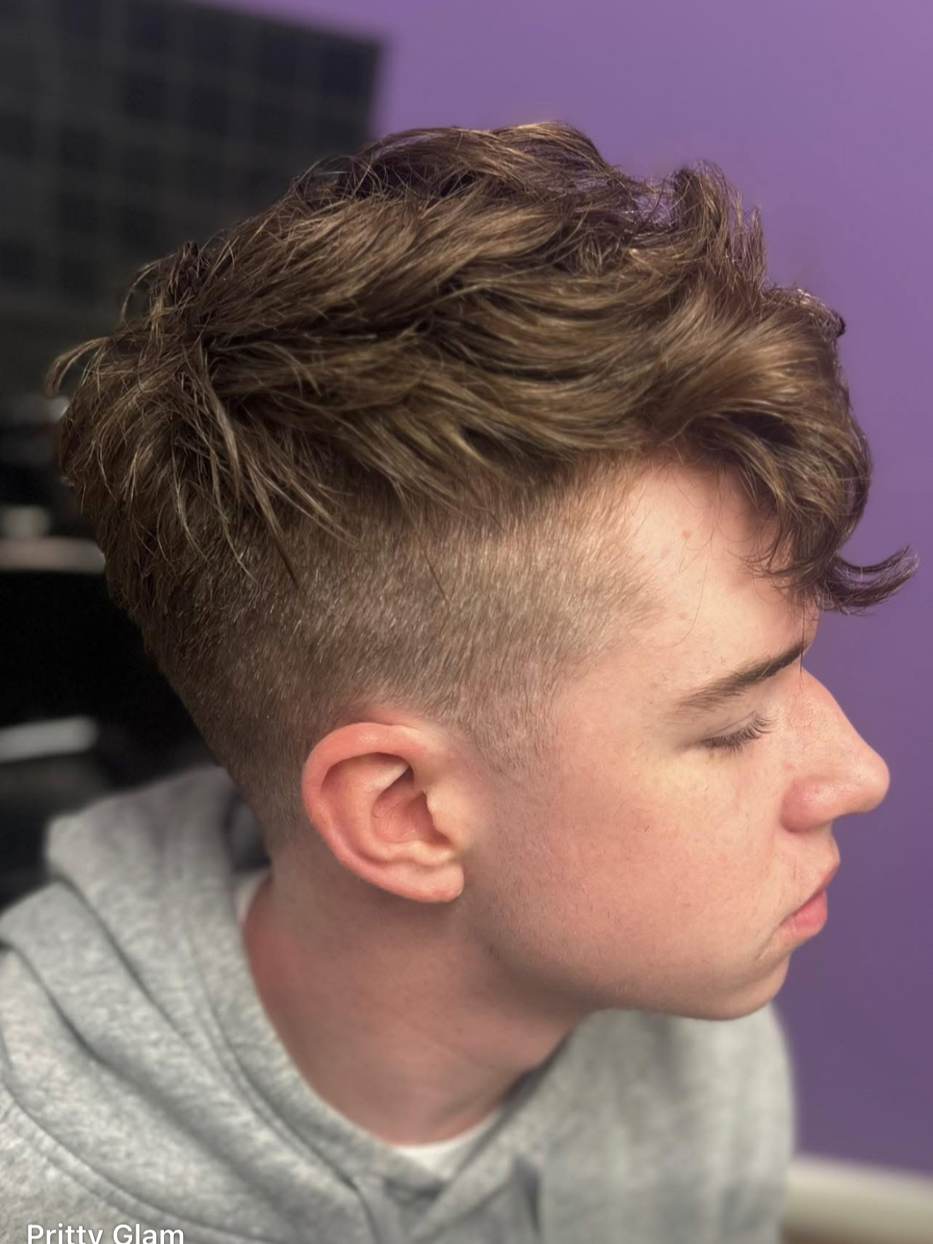 Close-up side profile of a young man with a modern haircut featuring a fade on the sides and longer, tousled hair on top, wearing a gray hoodie against a purple wall.