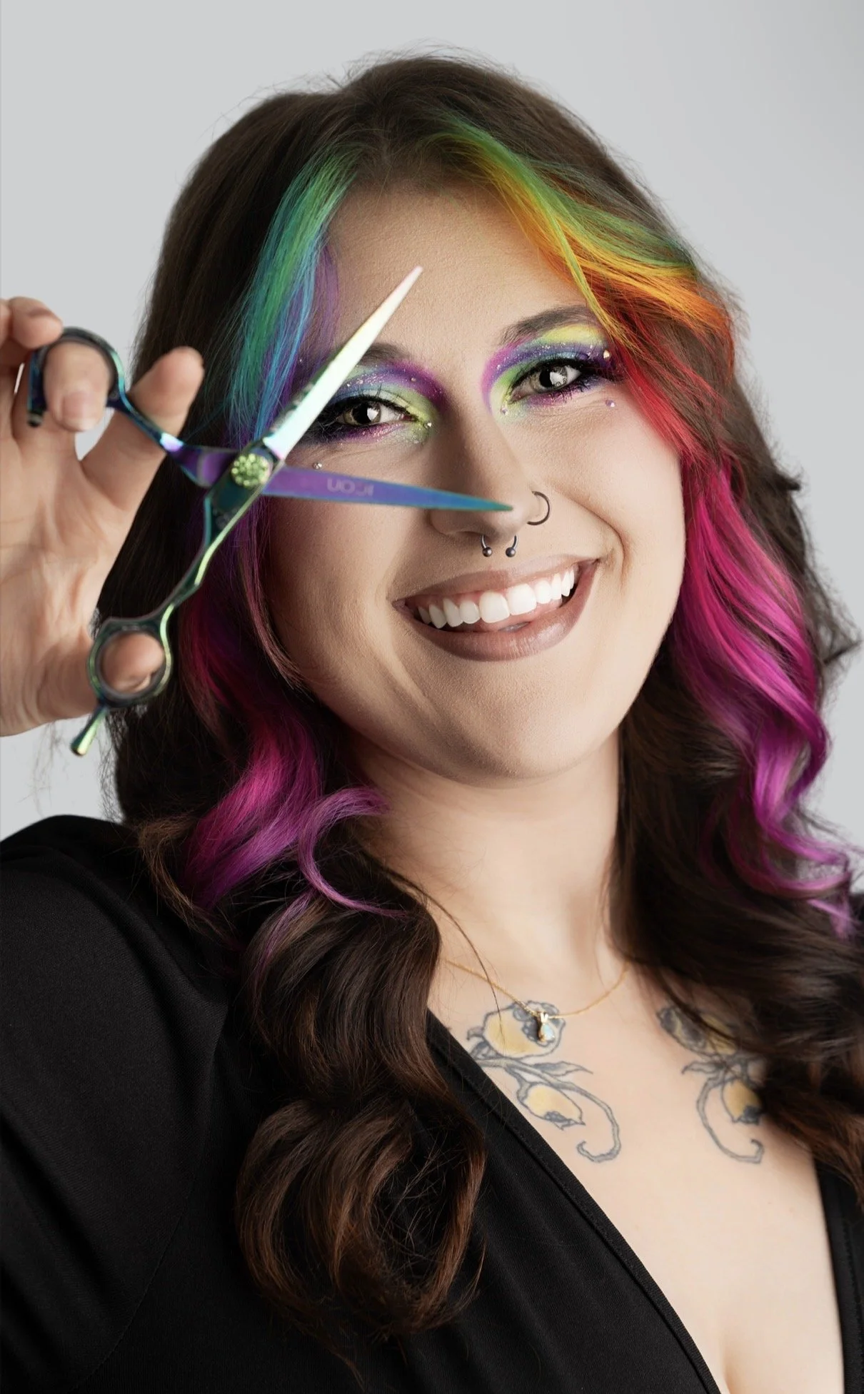 A woman with rainbow-colored hair and vibrant makeup holding hair cutting scissors, smiling.