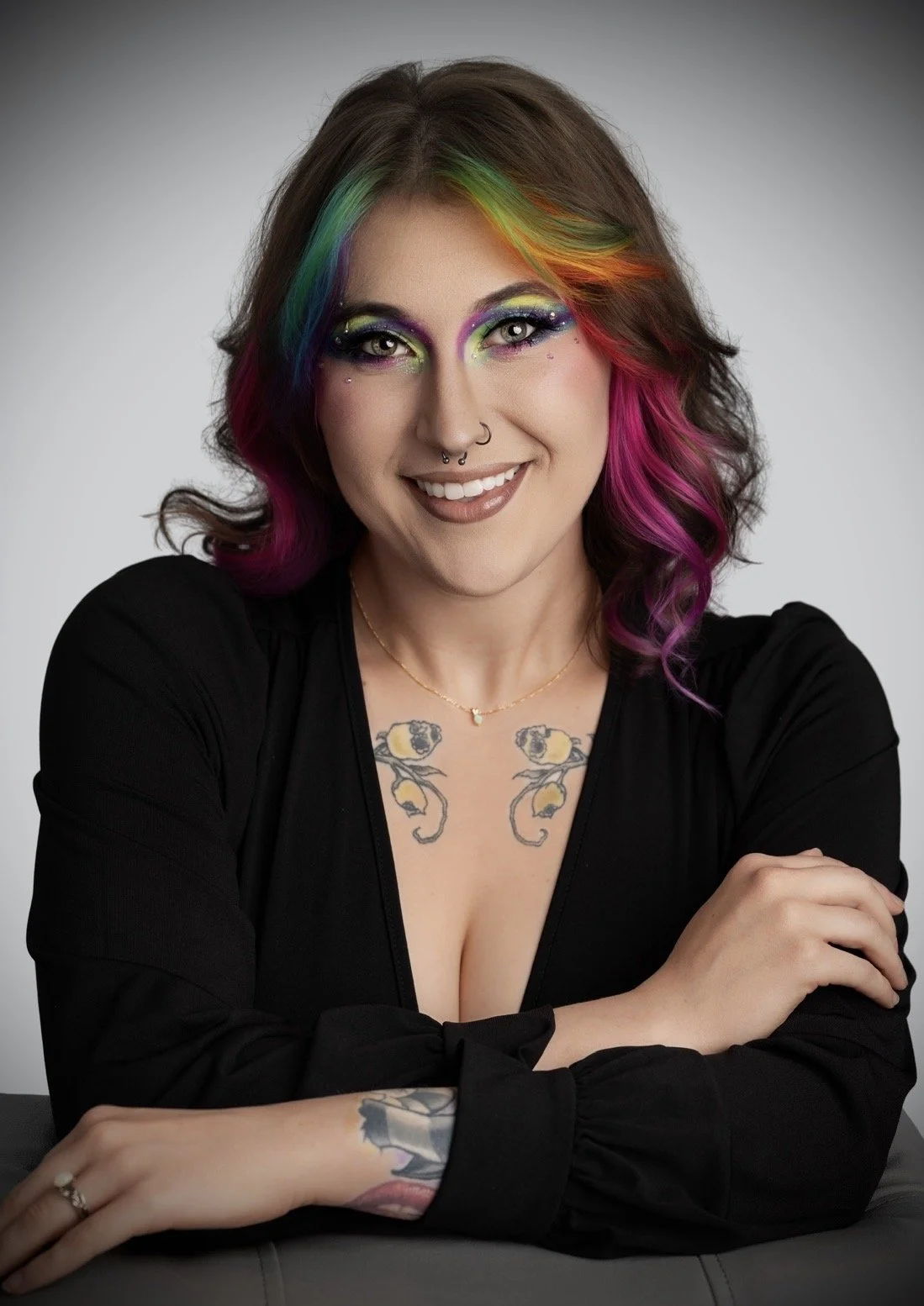 A woman with colorful rainbow hair and vibrant makeup, smiling with arms crossed, wearing a black top and displaying tattoos on her chest and arms.
