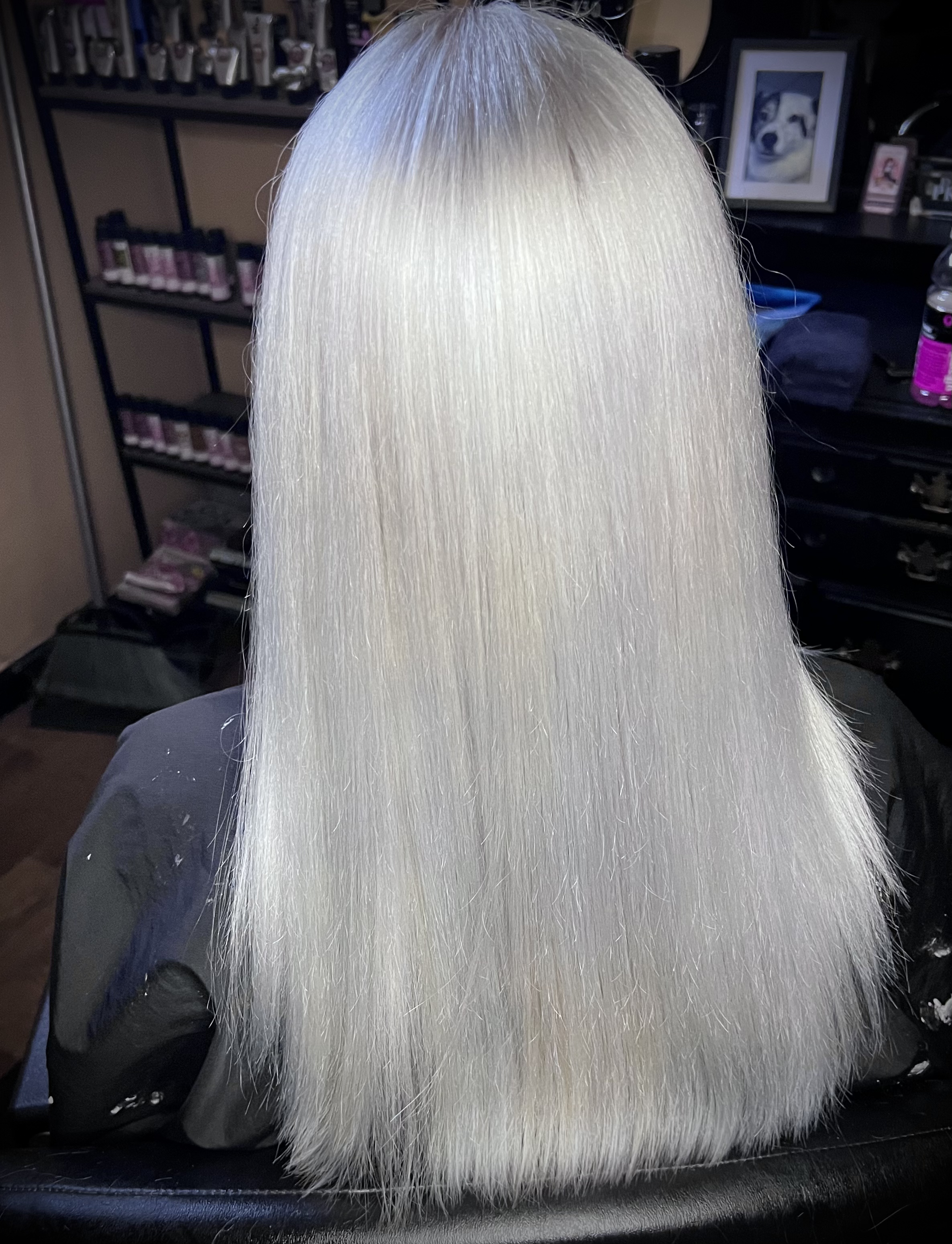 Close-up of platinum blonde hair with a smooth, straight texture, set against a background of beauty products and framed photos.