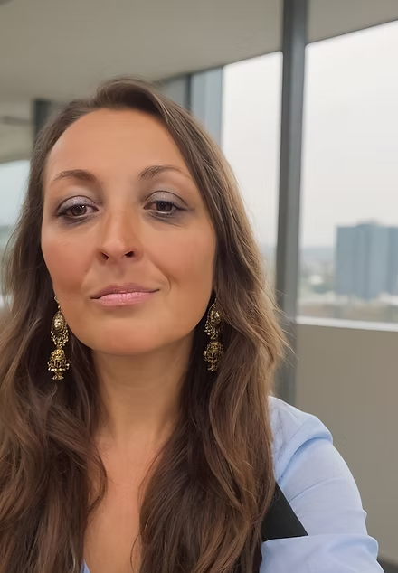 Close-up of a woman with long brown hair wearing gold earrings and a light blue shirt, inside a modern building with large windows and cityscape background.