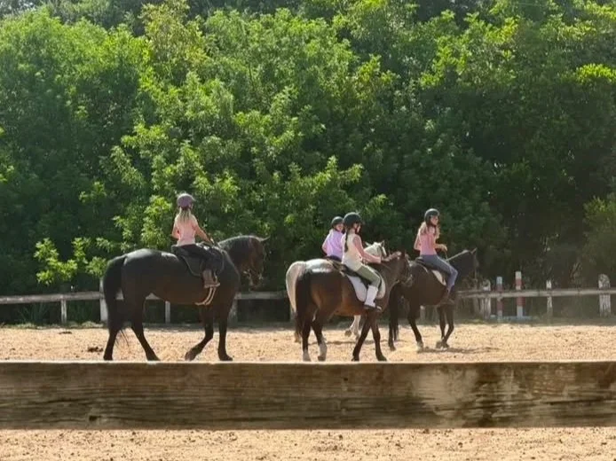 Four children riding horses in an outdoor riding arena with green trees in the background.